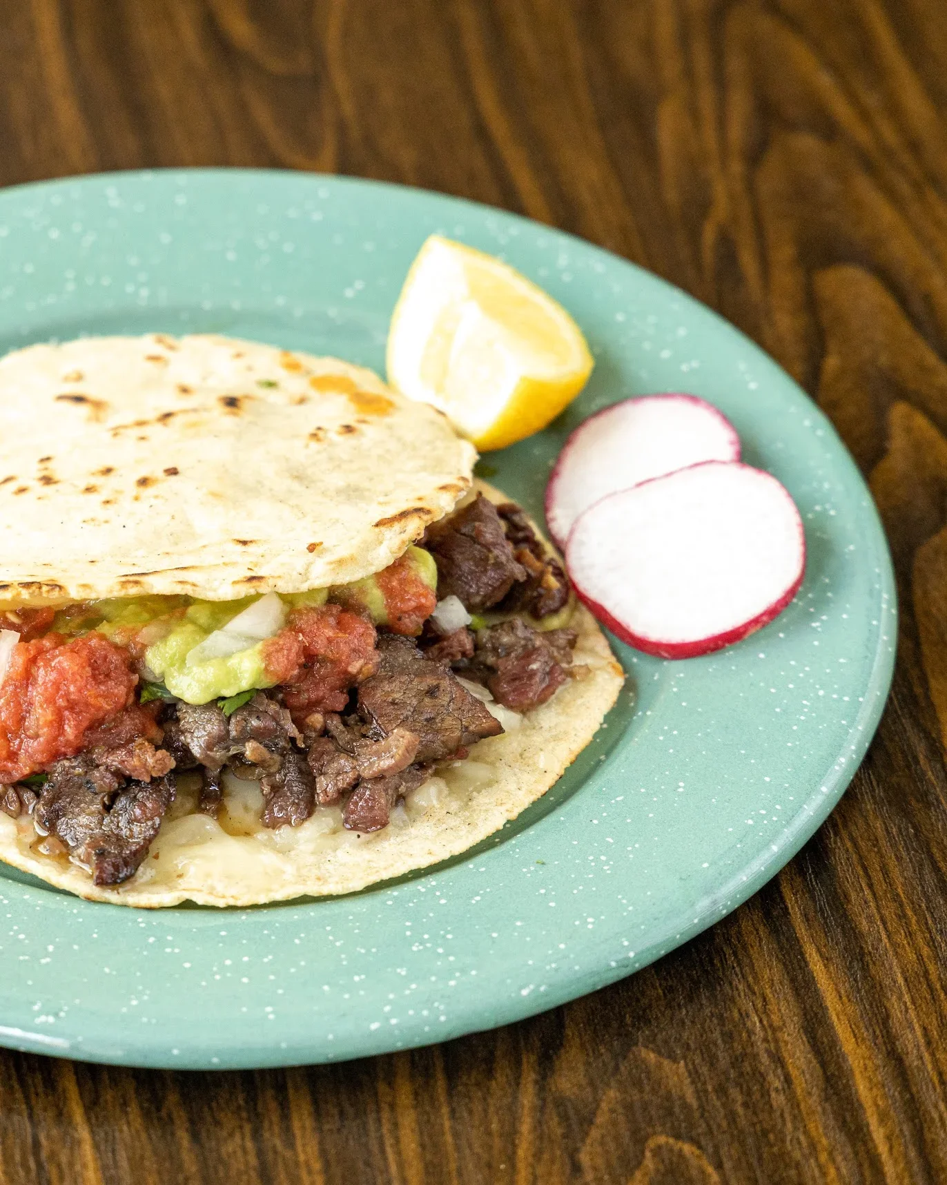 A plate with a taco filled with beef, guacamole, and salsa, a lemon wedge, and sliced radishes on a wooden table.