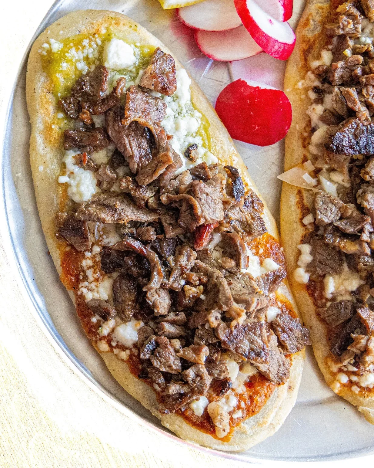 Steak and cheese sandwich on a bun with sliced radishes and tomato slices on the side.