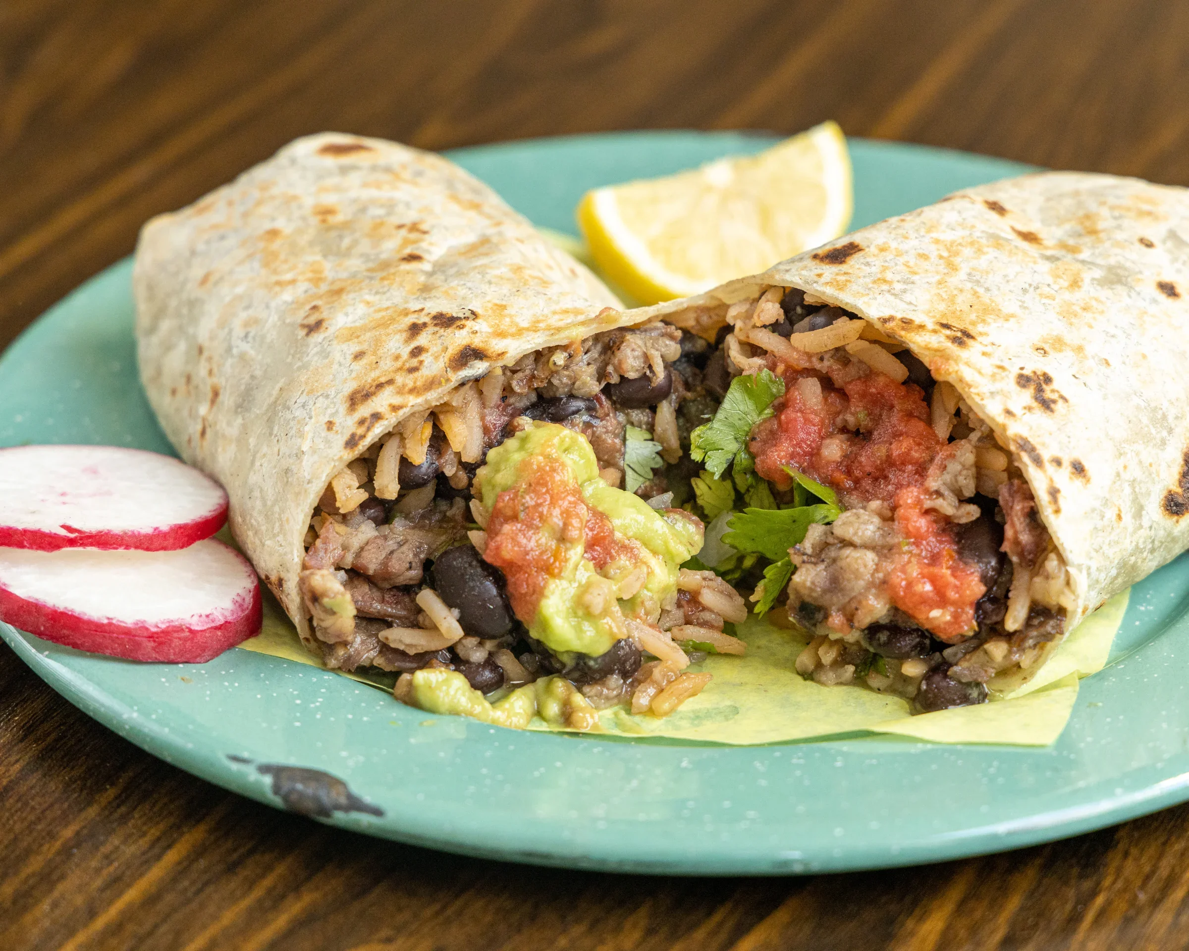 Close-up of a burrito cut in half on a green plate, with radish slices, lemon wedge, and salsa.