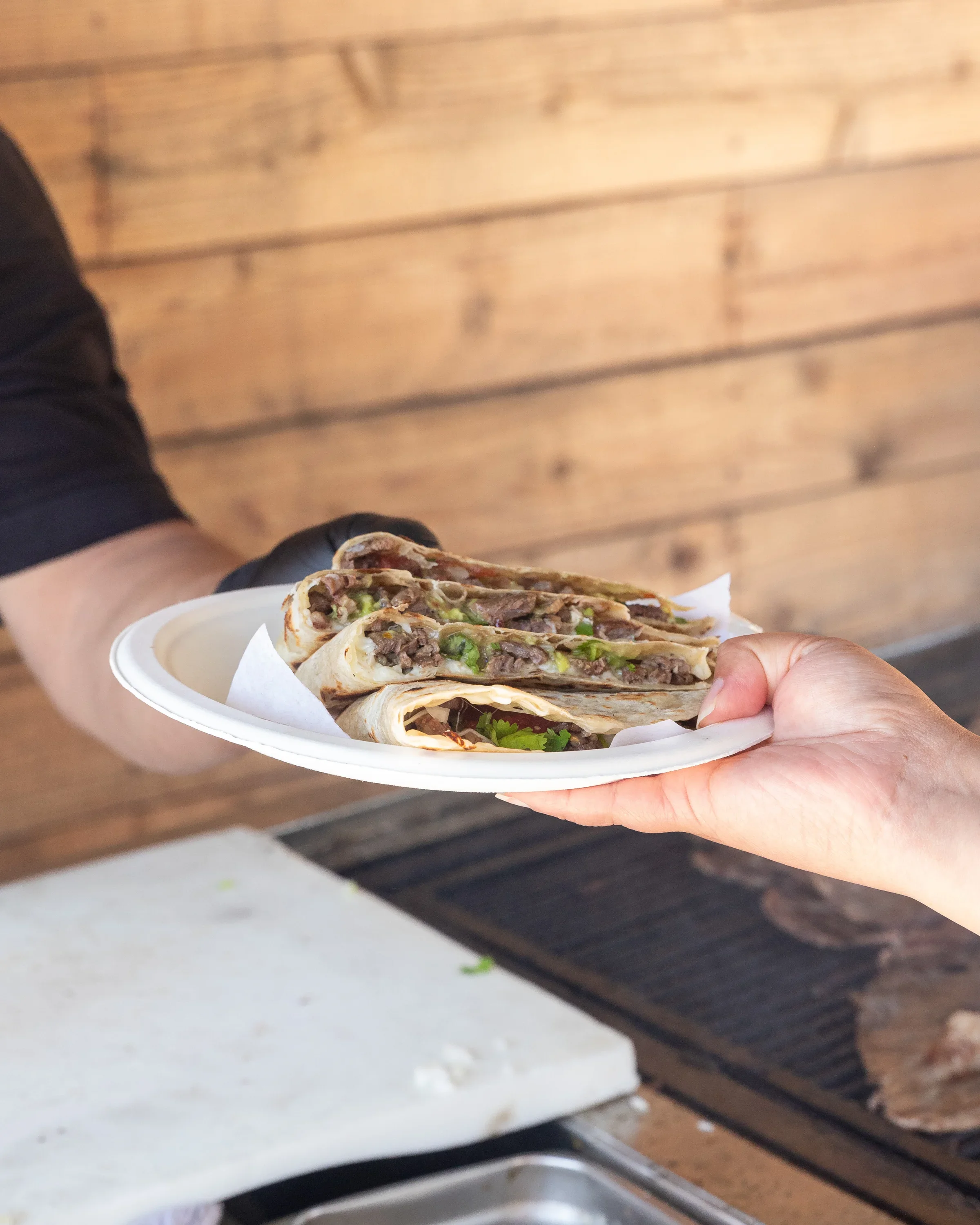 A person holding a white disposable plate with three folded steak and green pepper burritos, garnished with cilantro, against a wooden wall background.