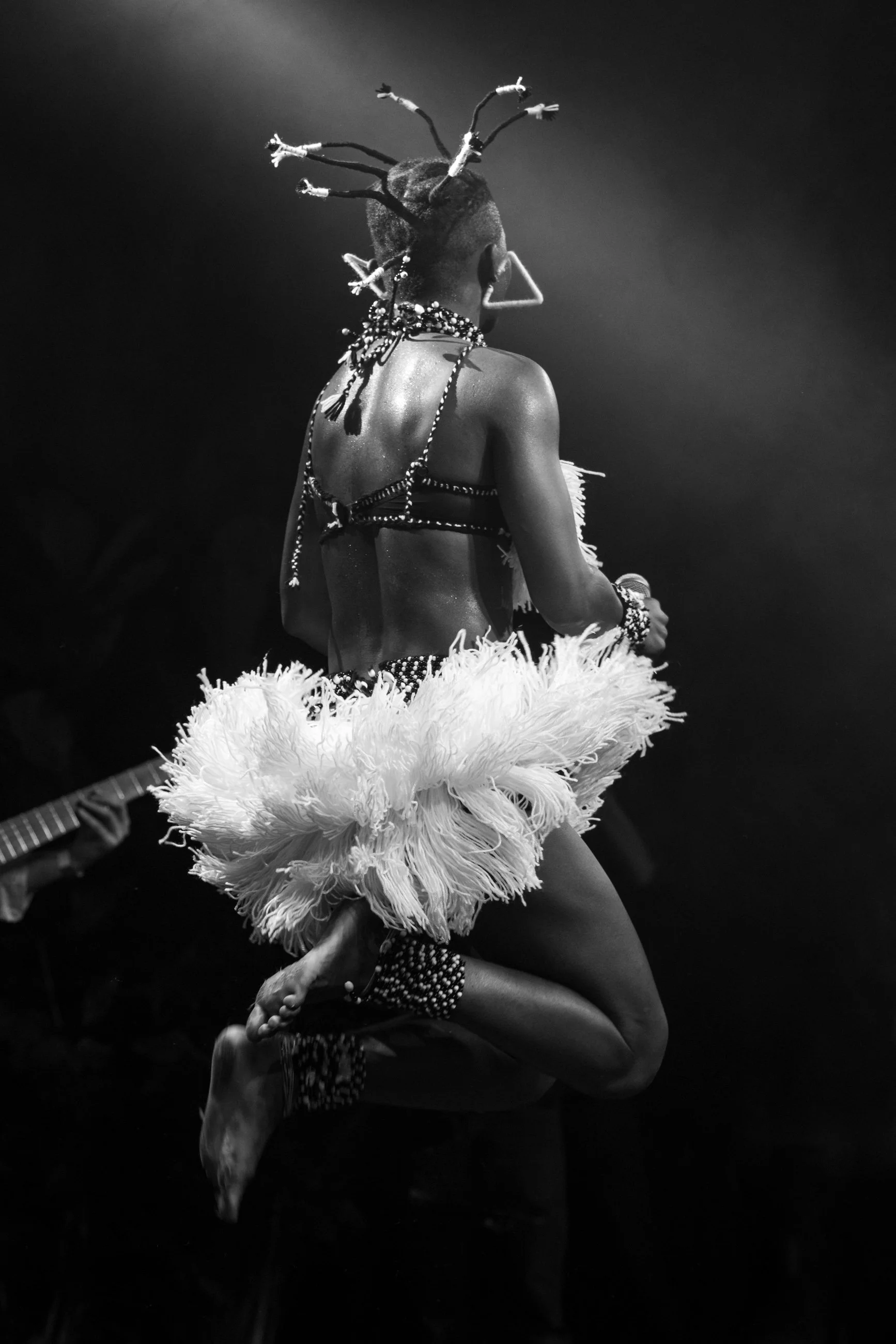 A performer dressed in traditional African attire, with beads and a feathered skirt, jumping or dancing on stage, holding a microphone, and seen from behind in black and white.