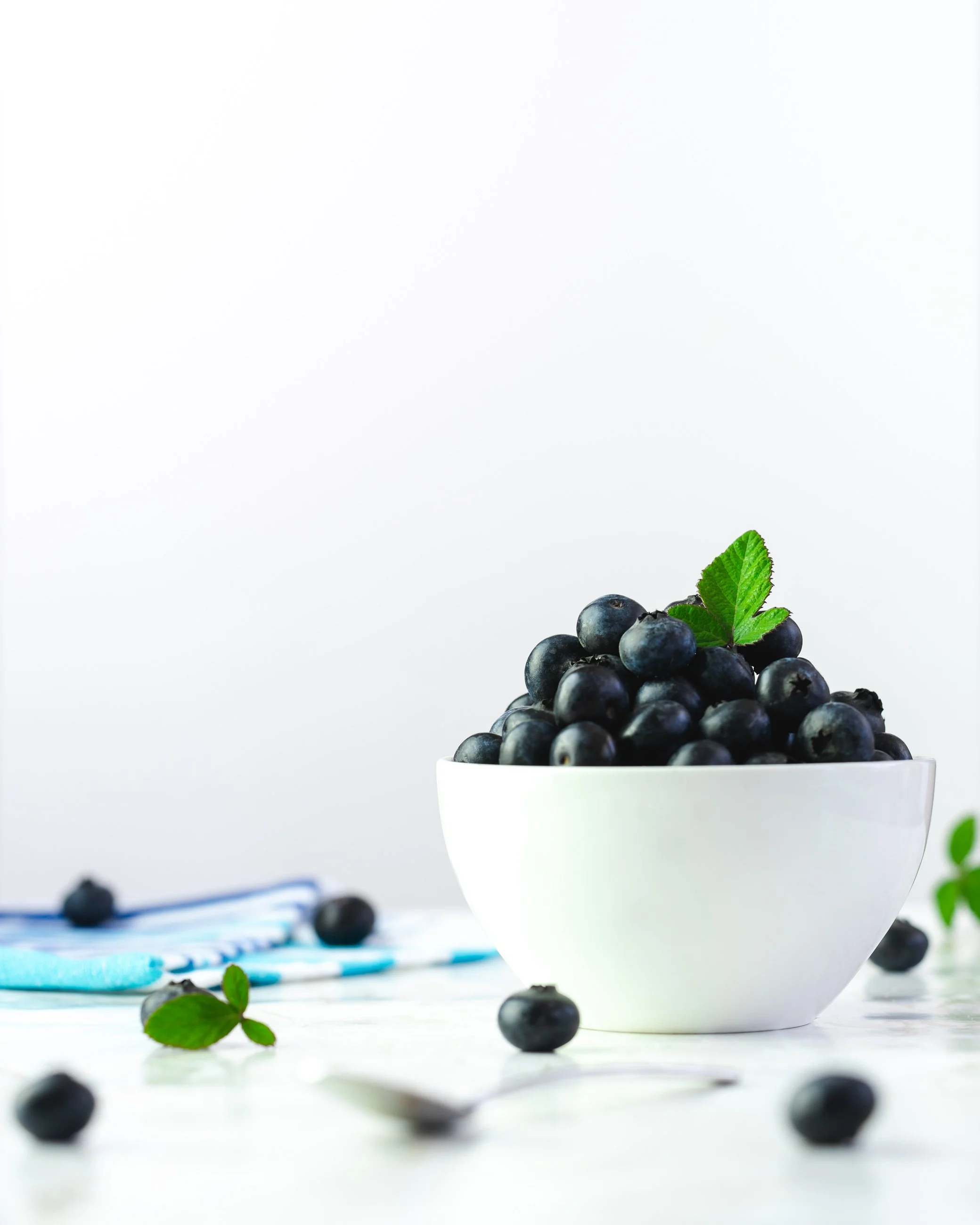 A white bowl filled with blueberries, topped with a green mint leaf, on a white surface with additional blueberries scattered around.