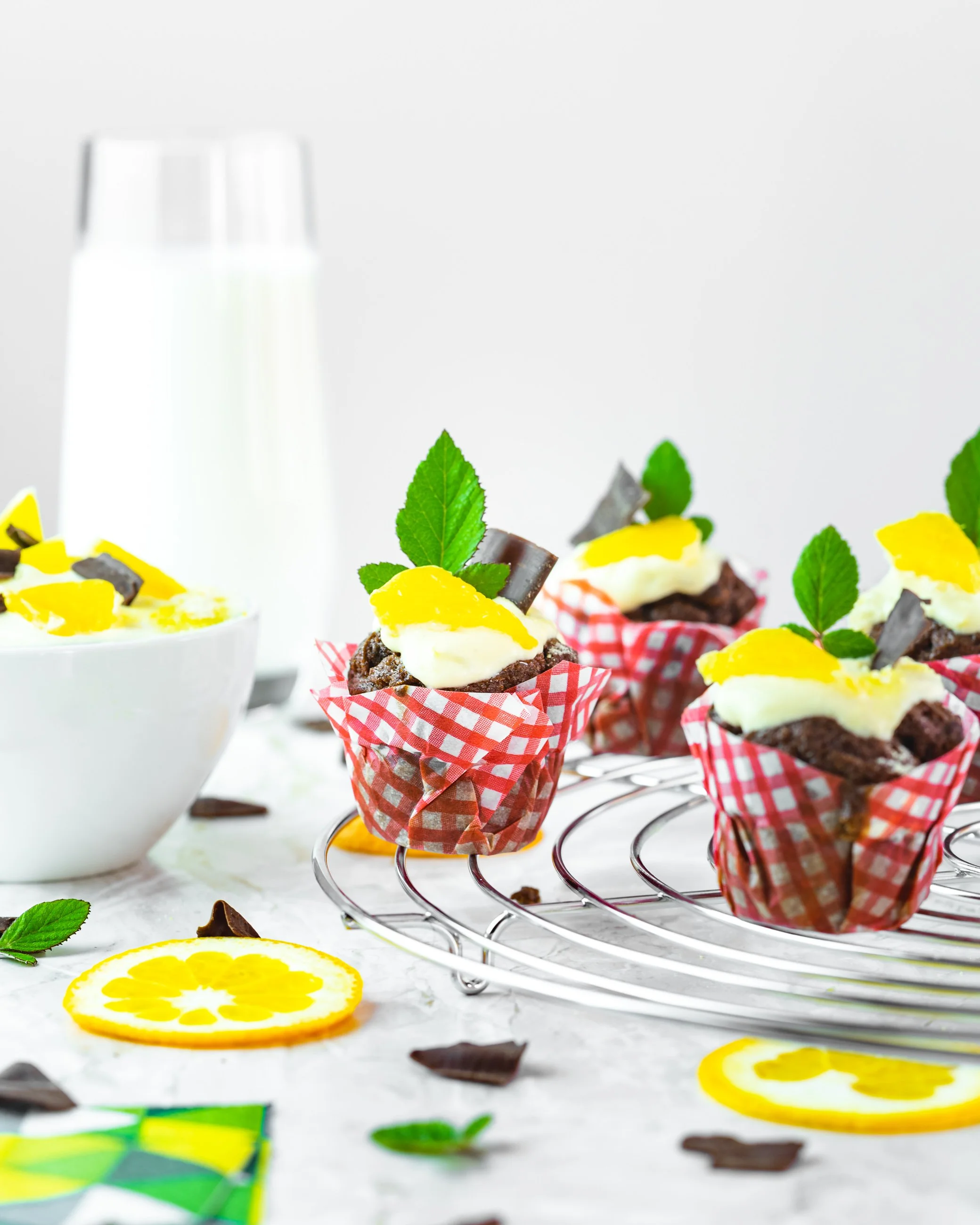 Chocolate cupcakes with white icing, yellow fruit topping, chocolate pieces, and mint leaves in red checkered paper liners on a cooling rack, with lemon slices and chocolate shards on a white surface, and a glass of milk in the background.