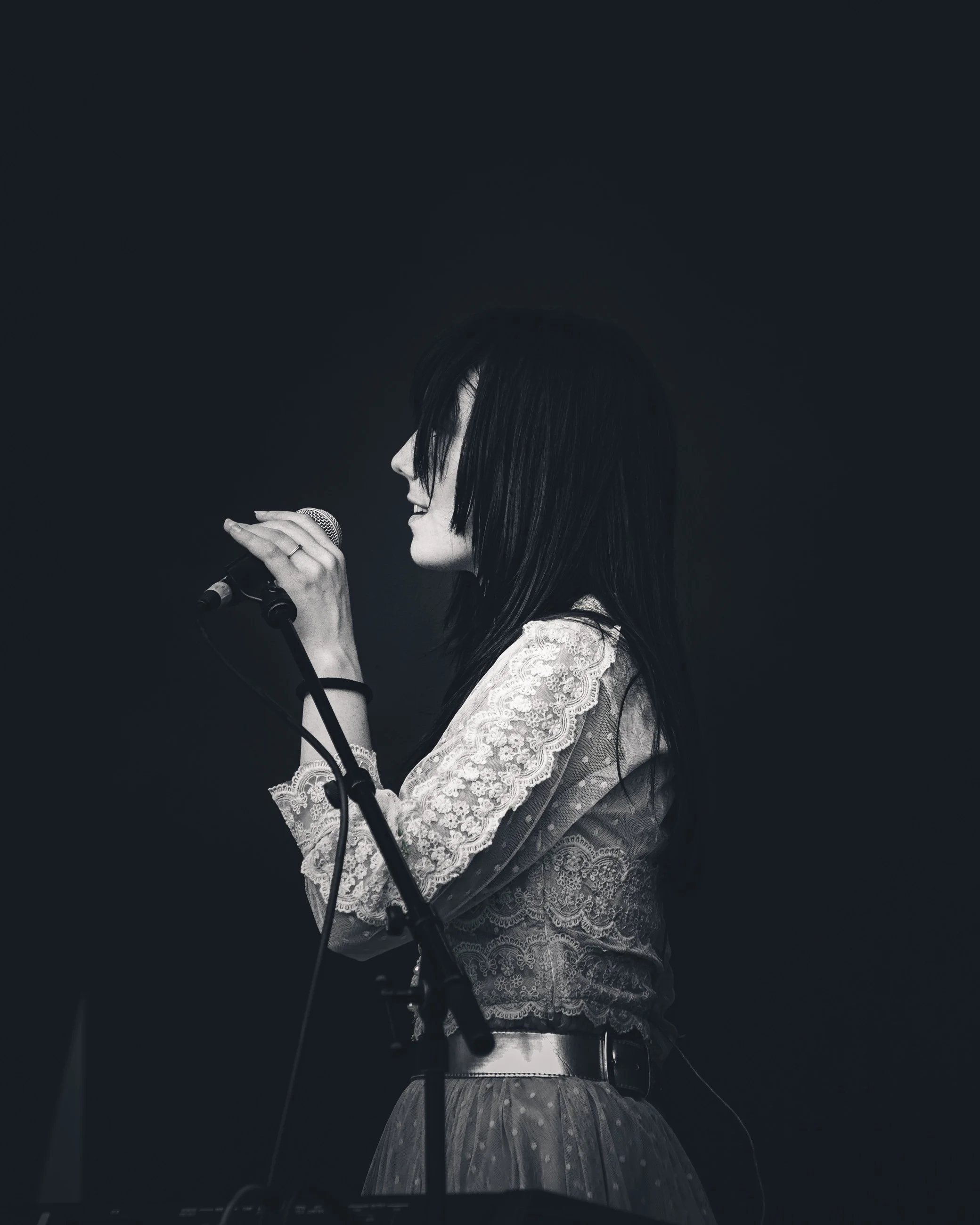 Black and white photo of a woman singing into a microphone on stage, wearing a lace top and a skirt, with dark hair.
