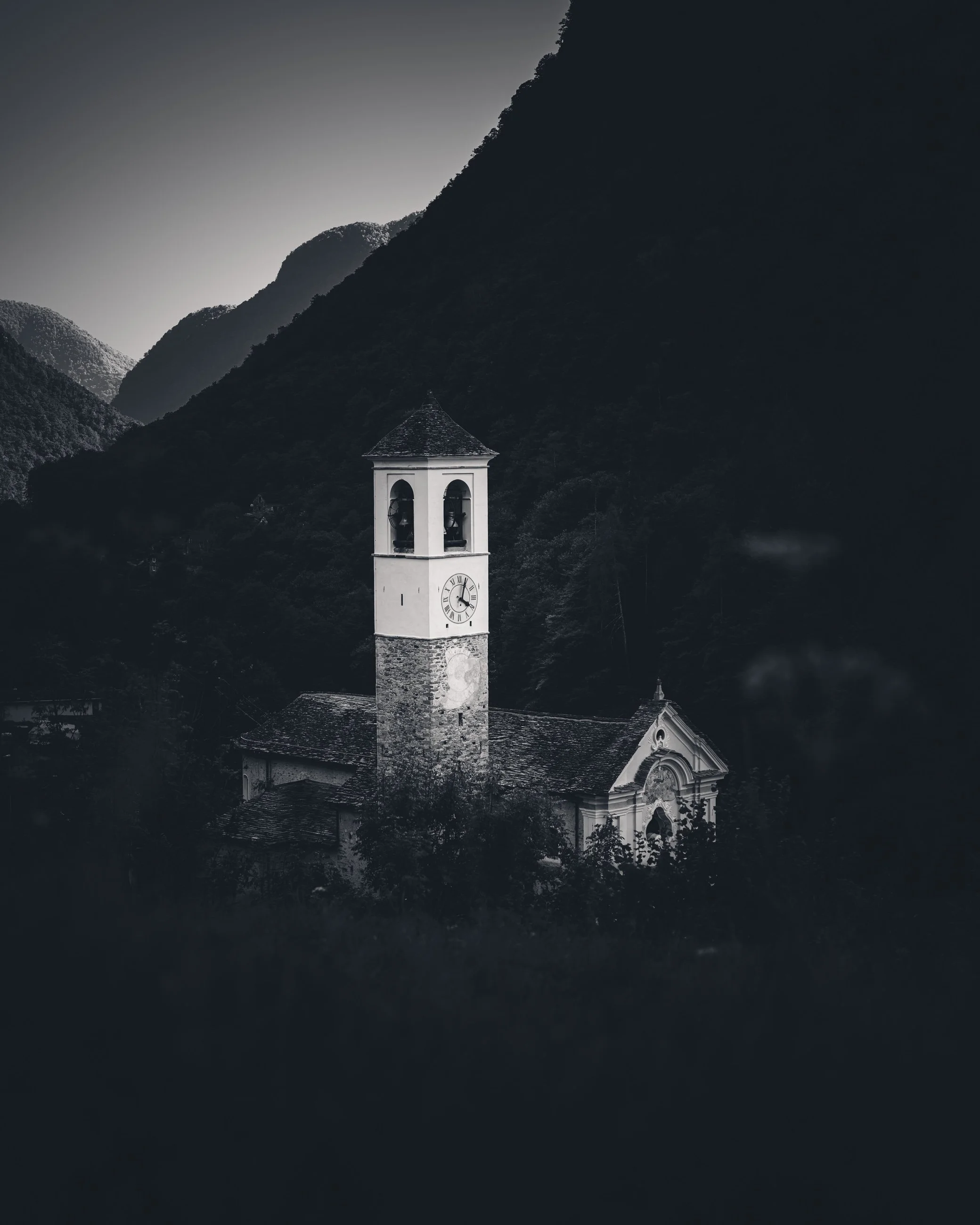 Black and white photo of a church with a clock tower, surrounded by tall mountains and trees.