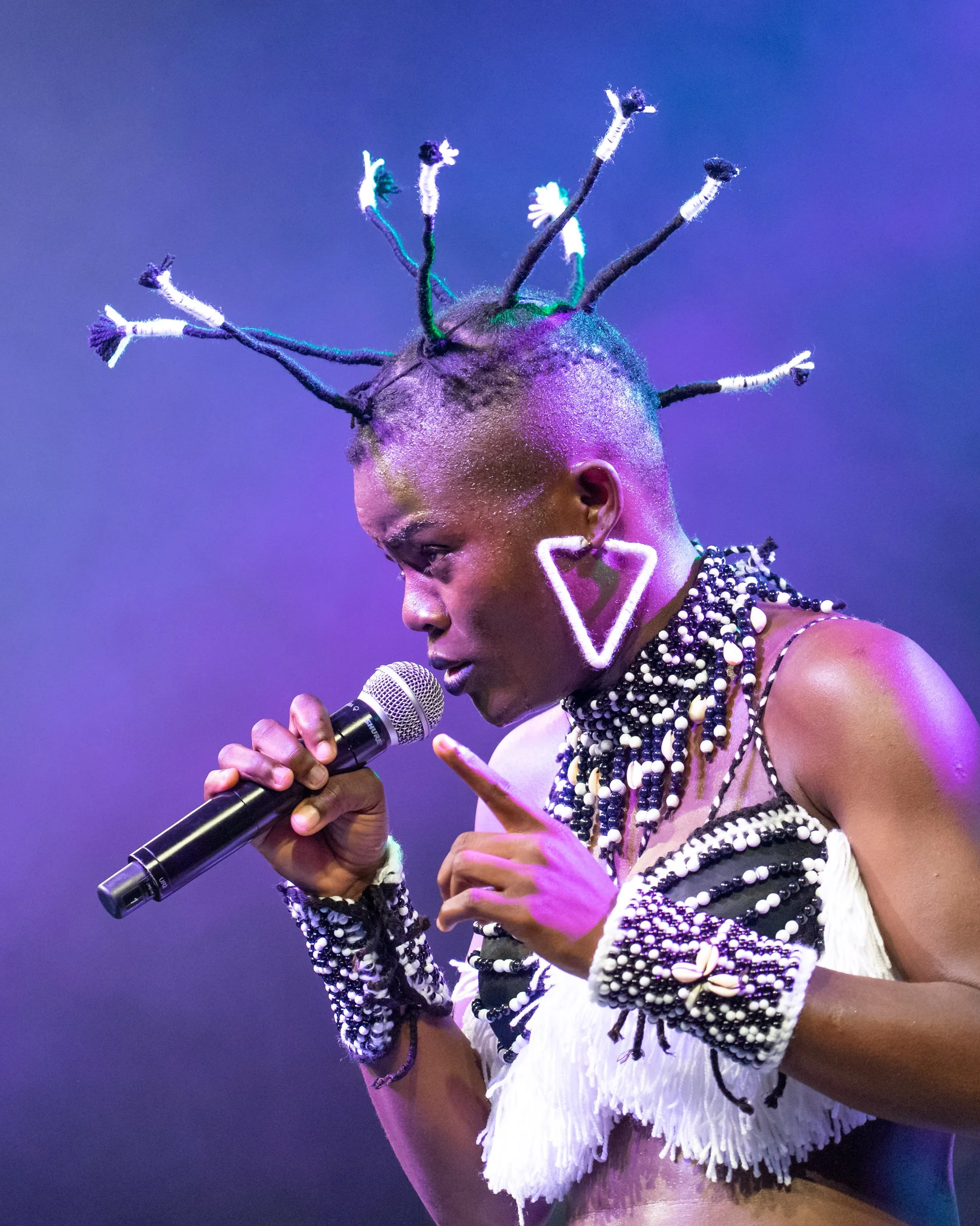 A performer singing into a microphone, wearing traditional tribal attire with bead necklaces, bracelets, and large heart-shaped earrings, and with hair styled in multiple dreadlocks decorated with flowers and beads.