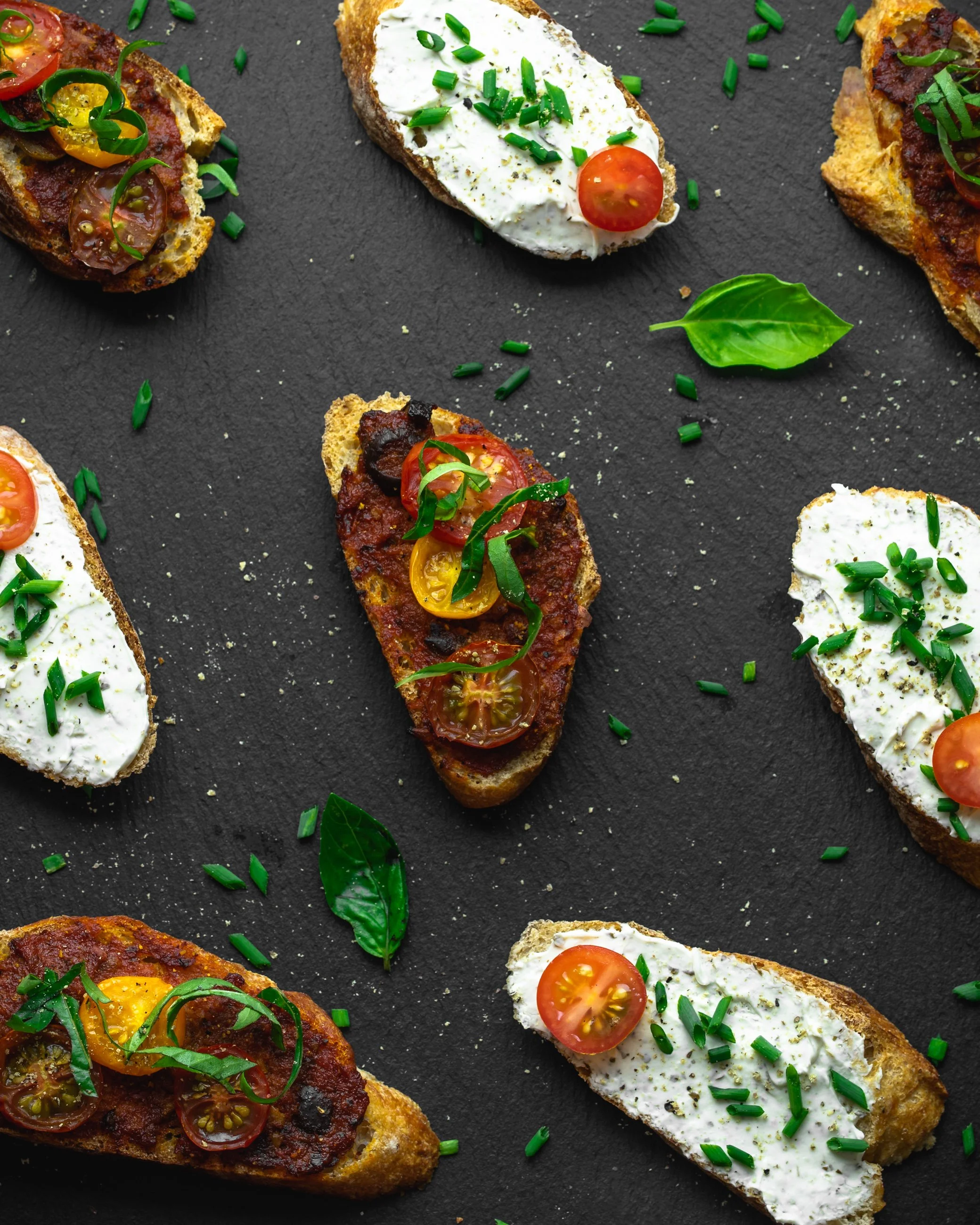 Assorted toasted bread slices topped with cherry tomatoes, fresh herbs, and cheese, arranged on a dark slate surface.