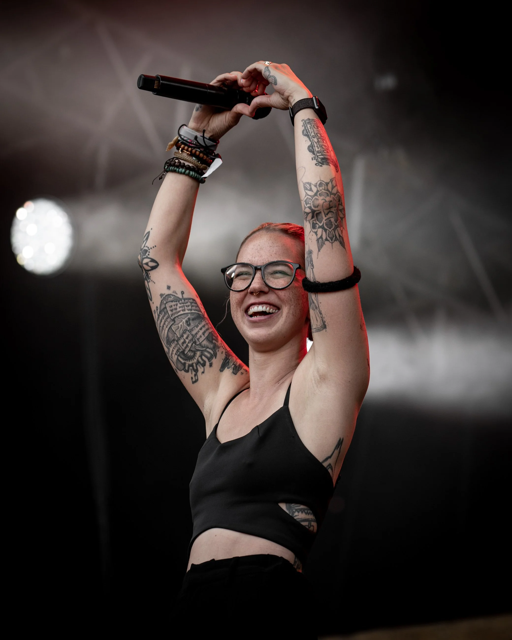 A woman with tattoos and glasses is smiling and holding a microphone up in the air at a concert or event.