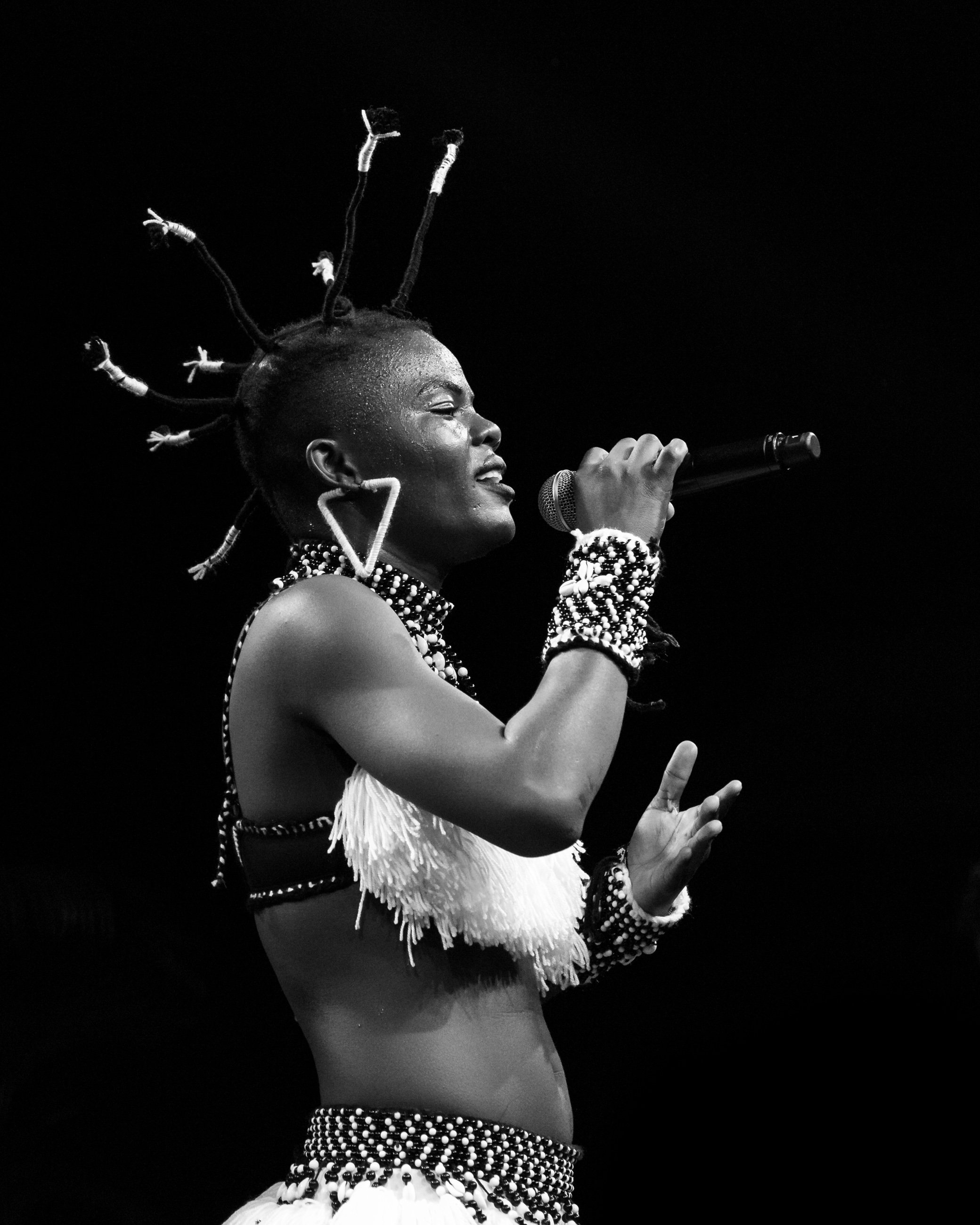 A black and white photo of a woman with styled hair and multiple earrings, singing into a microphone, wearing traditional African attire decorated with beads and fringe.