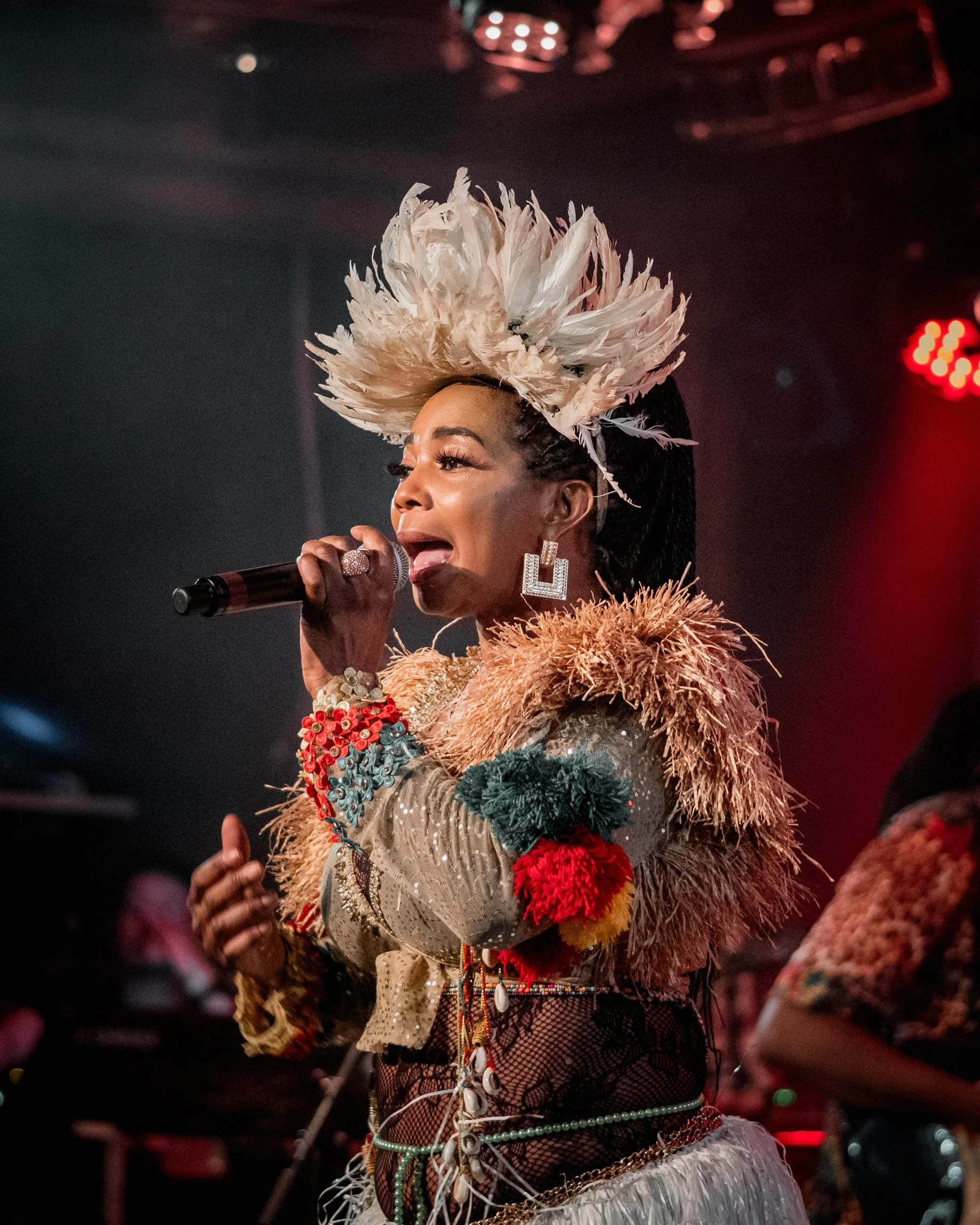 A woman singing into a microphone on stage, wearing an elaborate feathered headpiece, large earrings, and a colorful outfit with feathers and bead decorations.