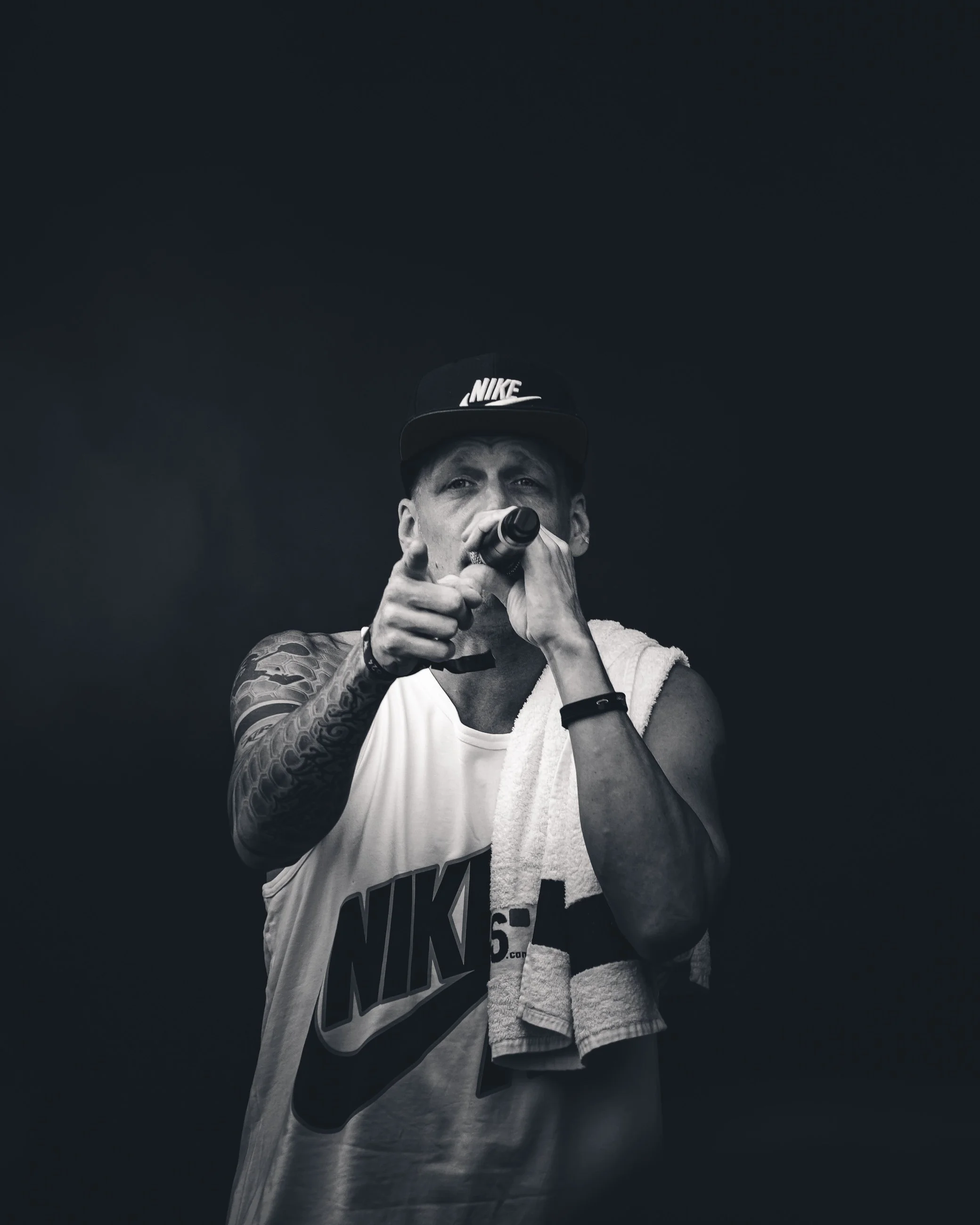 A man wearing a Nike tank top and a Nike cap is holding a microphone and pointing towards the camera, with a towel draped over his shoulder, against a dark background.
