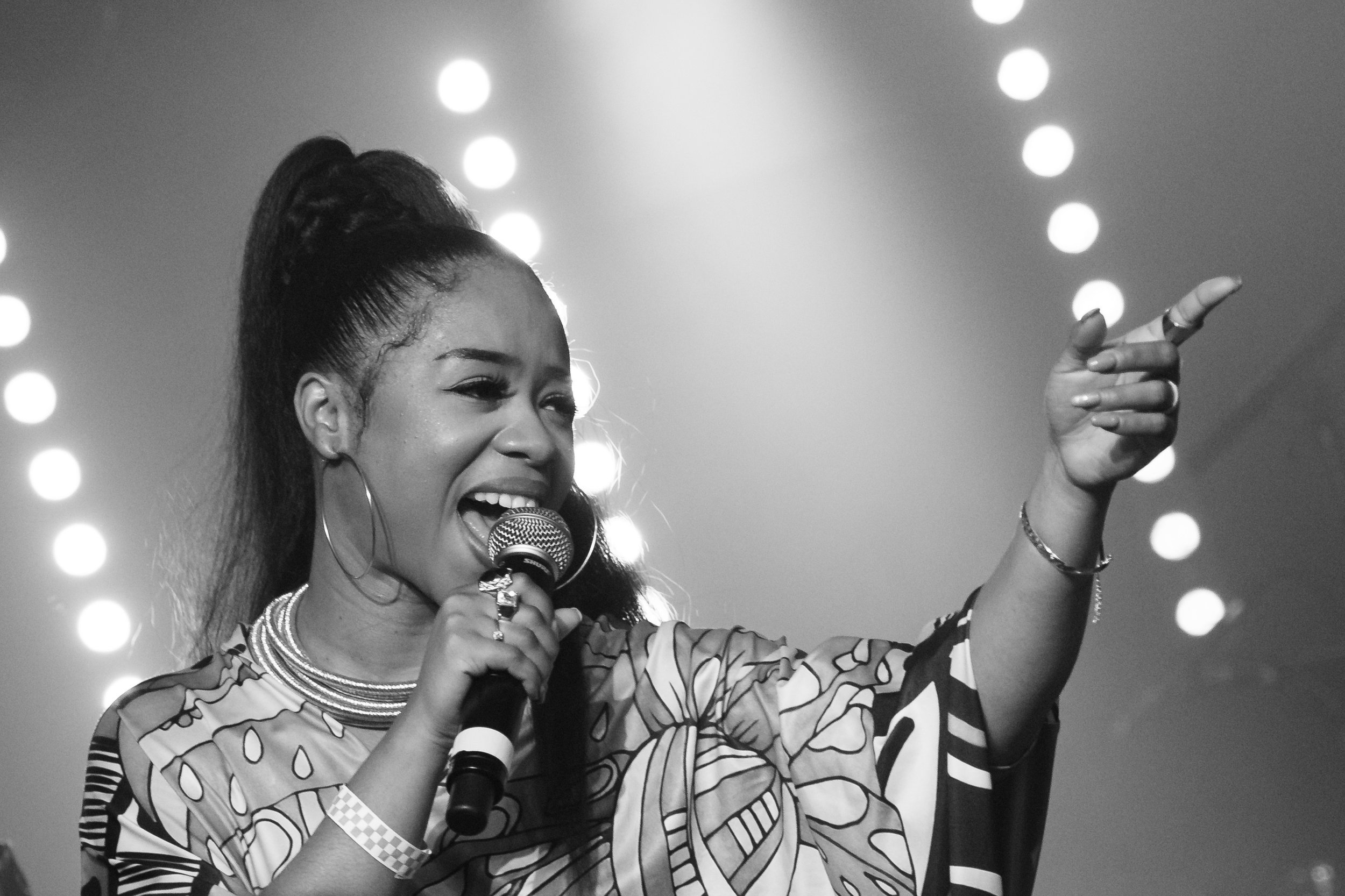 A woman singing passionately into a microphone on stage, with her right arm raised and finger pointing, against a background of stage lights.