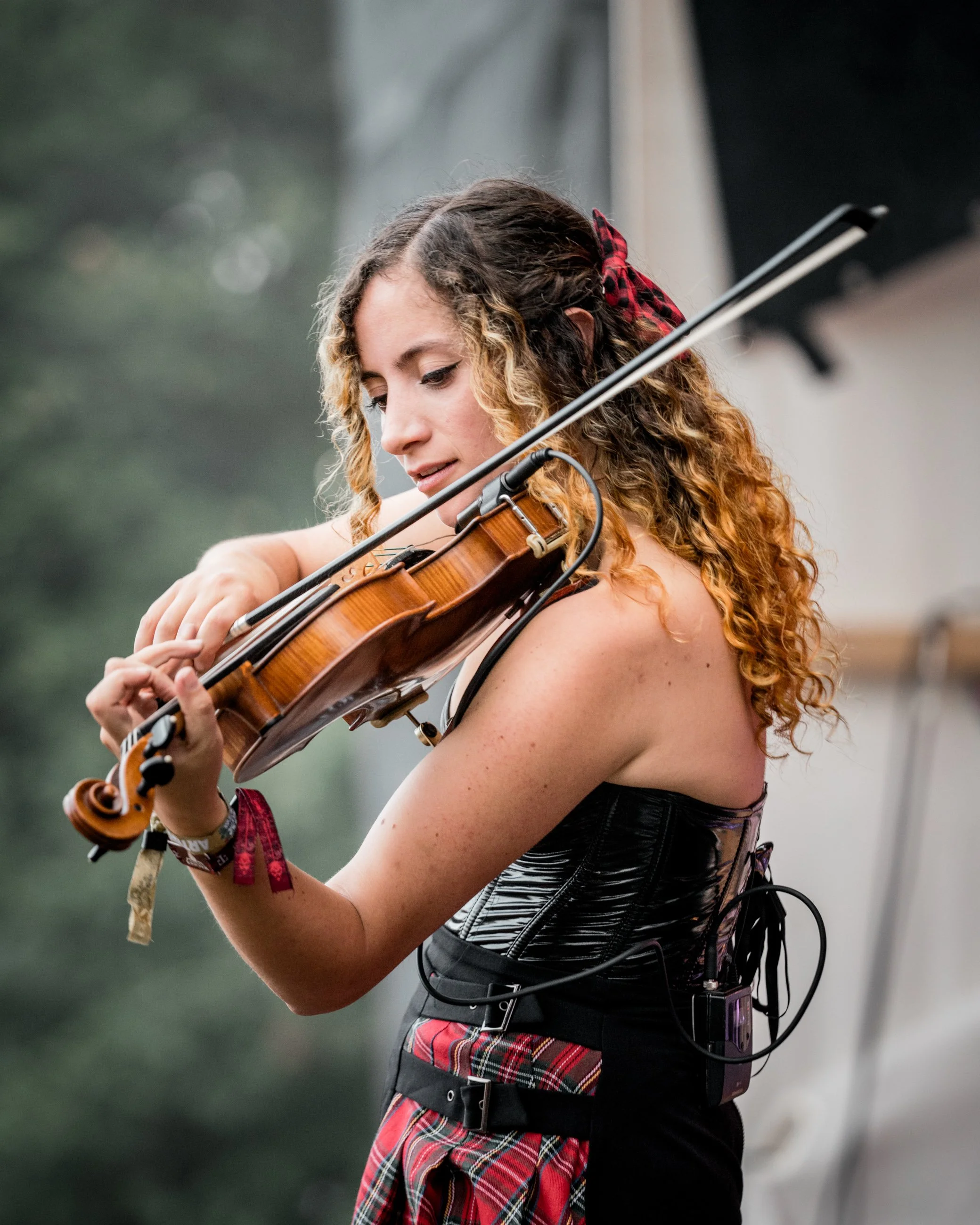 A woman playing a violin outdoors with a plaid shirt tied around her waist and colorful wristbands.