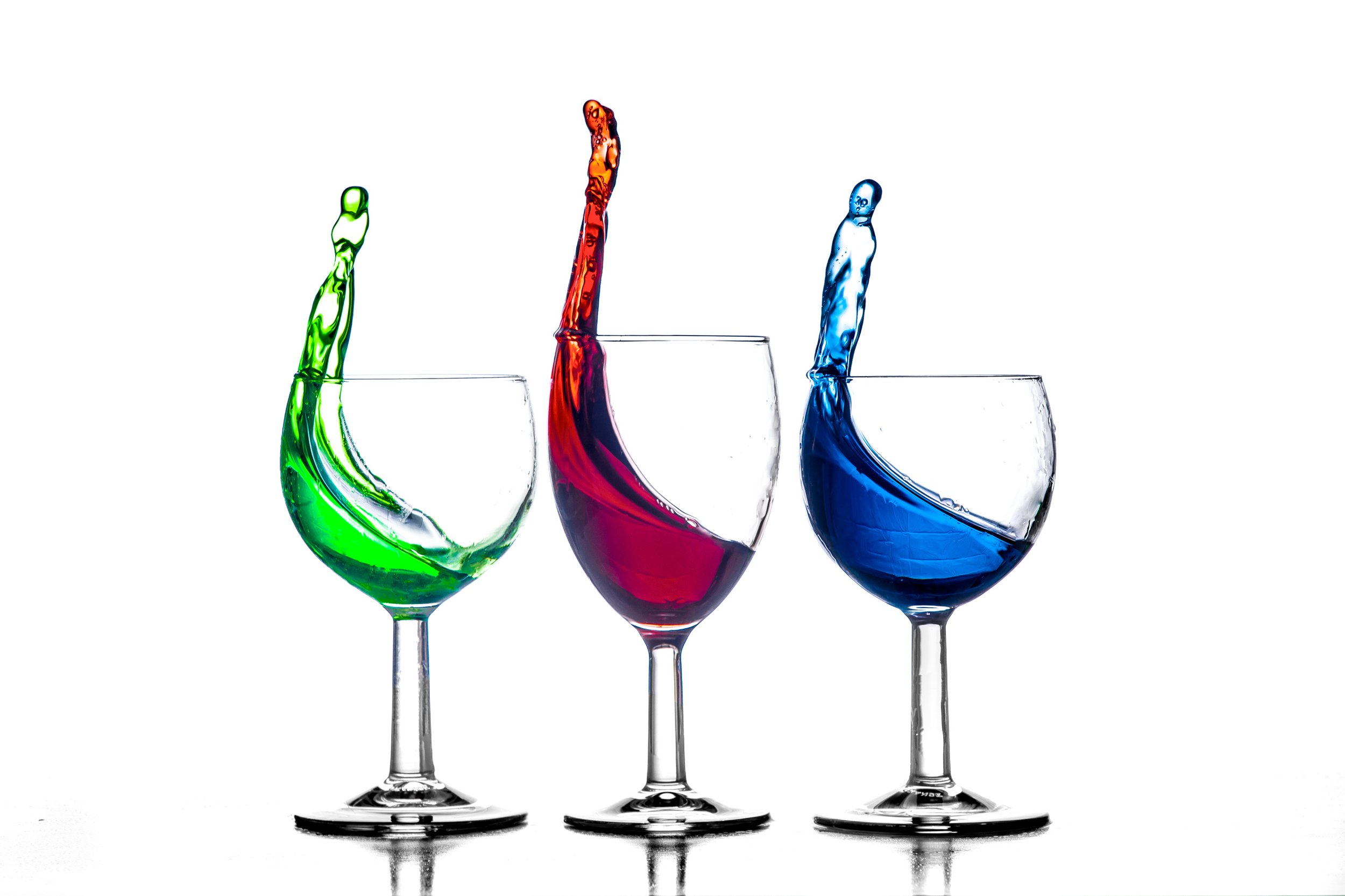 Three wine glasses with colored liquids (green, red, blue) splashing out of them against a white background.