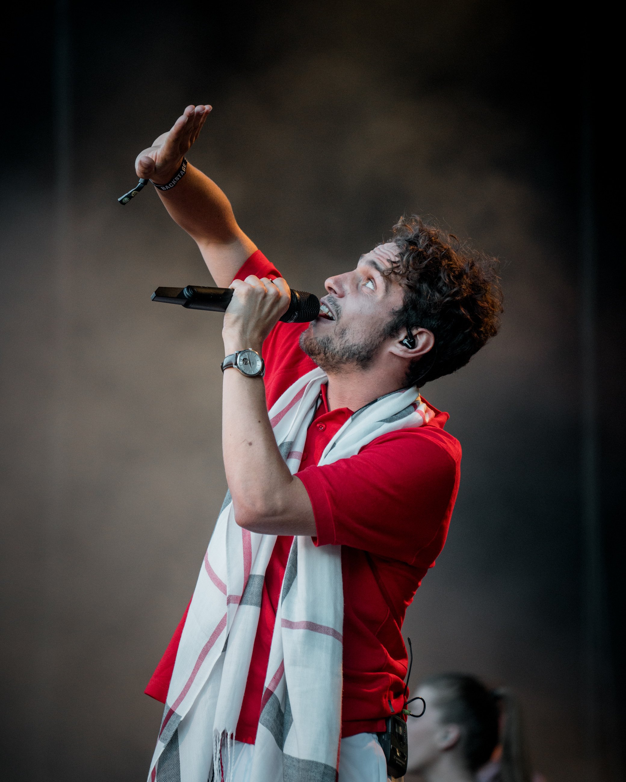 Male singer wearing a red shirt, holding a microphone, with a scarf around his neck, gesturing while performing on stage.