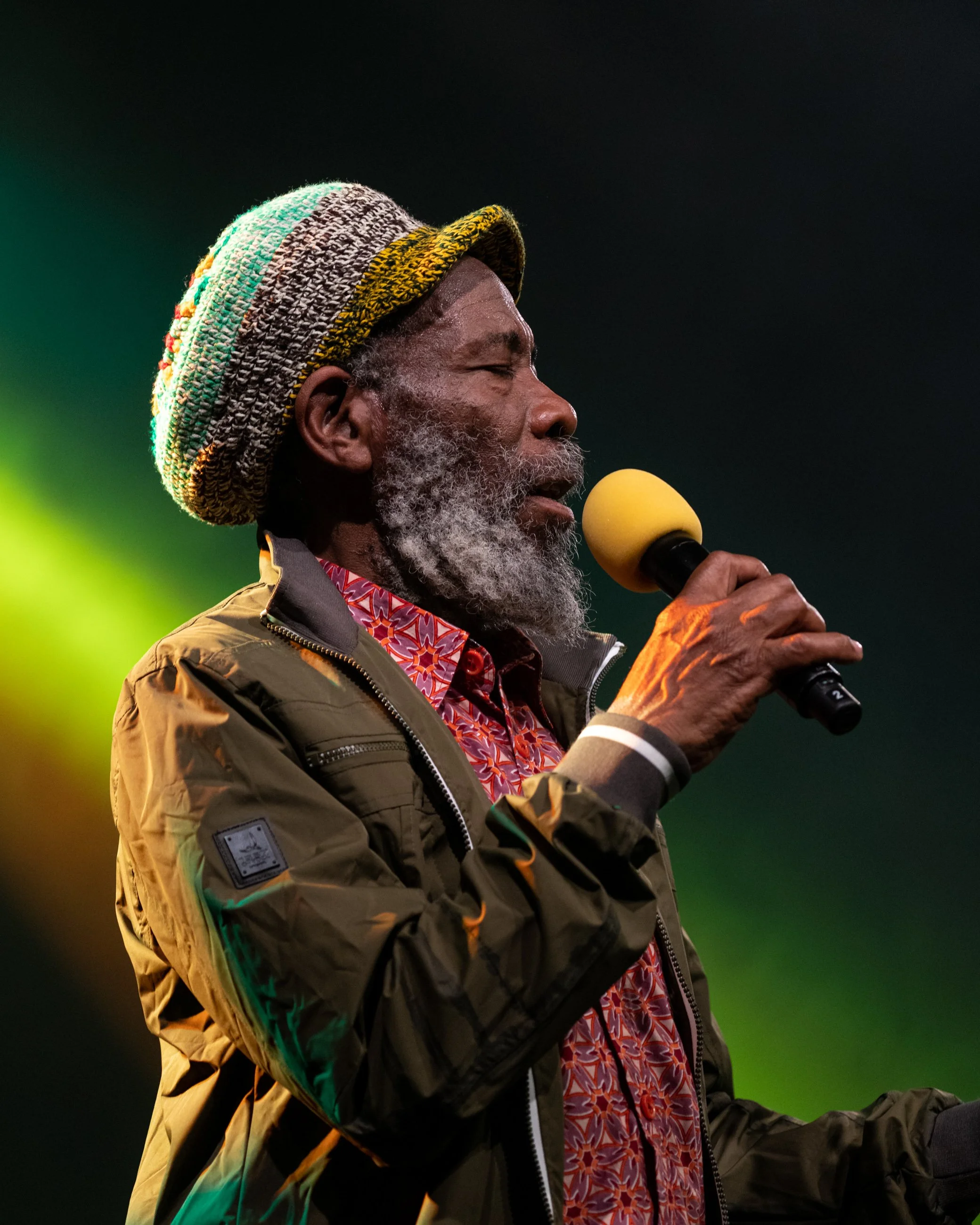 An elderly man with a grey beard and moustache, wearing a colorful knit hat, a patterned shirt, and a green jacket, holds a yellow microphone and appears to be speaking or singing.