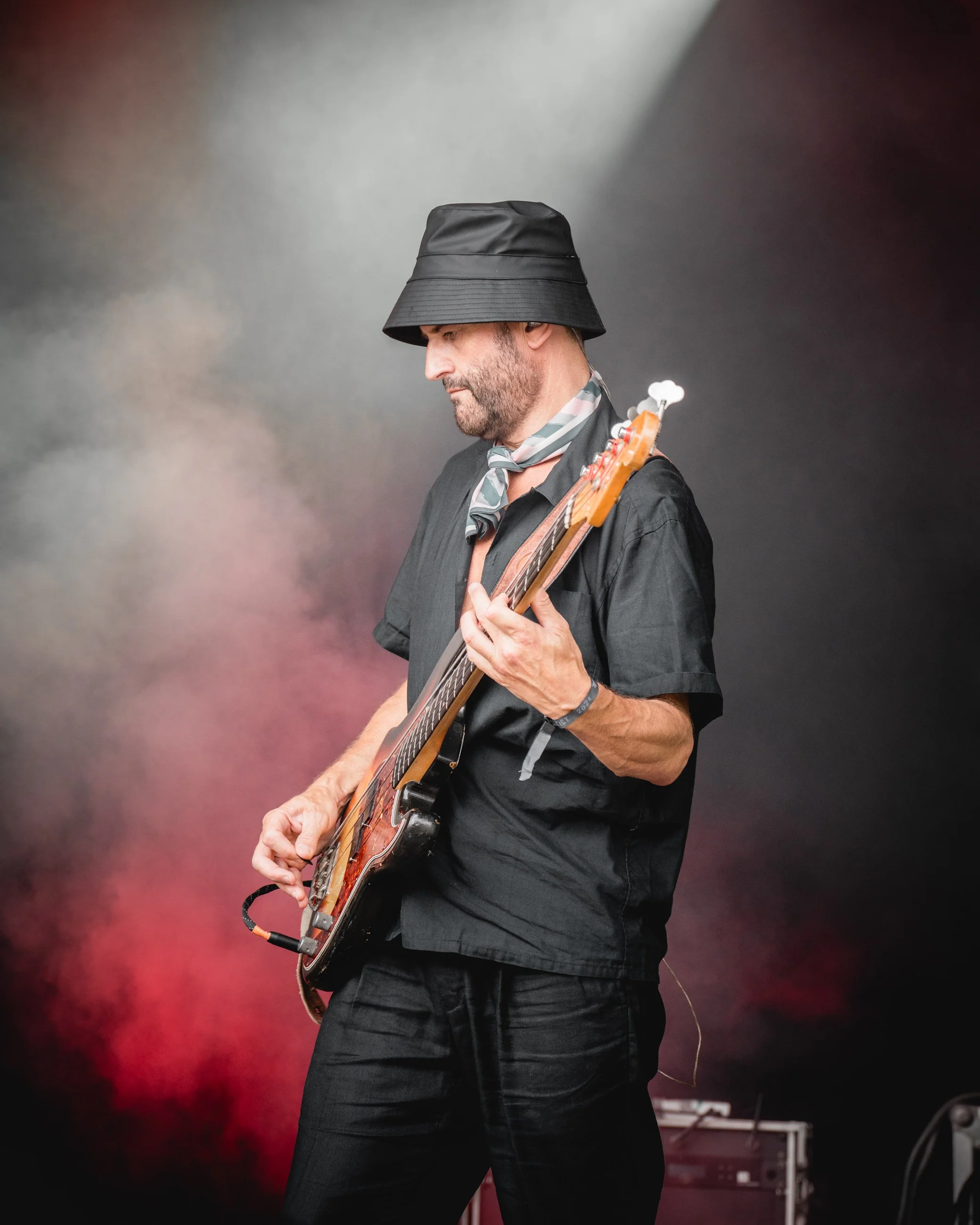 A man in a black hat, black shirt, and black pants playing an electric guitar on stage with colorful smoky background.
