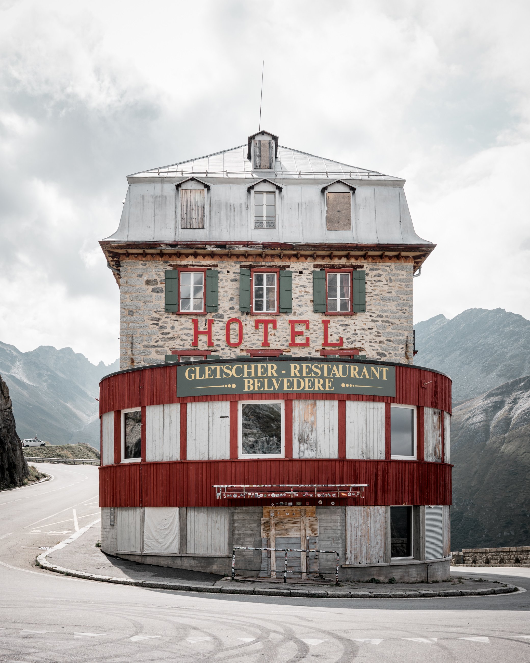 A rustic hotel building with green shutters, a stone facade, hezhigh windows, and a red and white wooden exterior, situated in mountainous terrain, with a sign for Gletscher-Restaurant Belvedere.