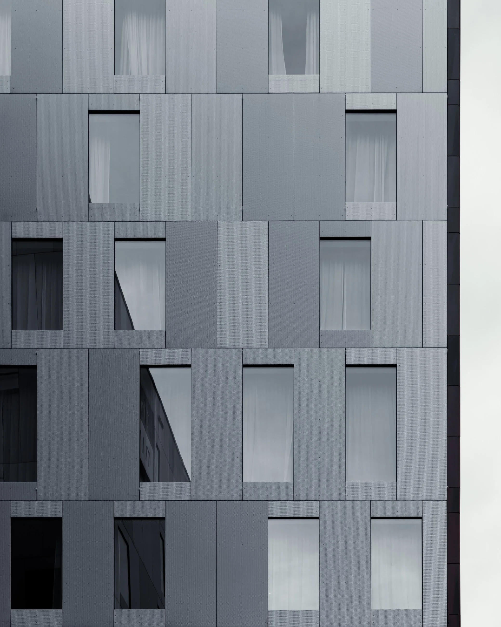 Close-up of a modern building's gray facade with multiple rectangular windows, some of which have curtains visible.