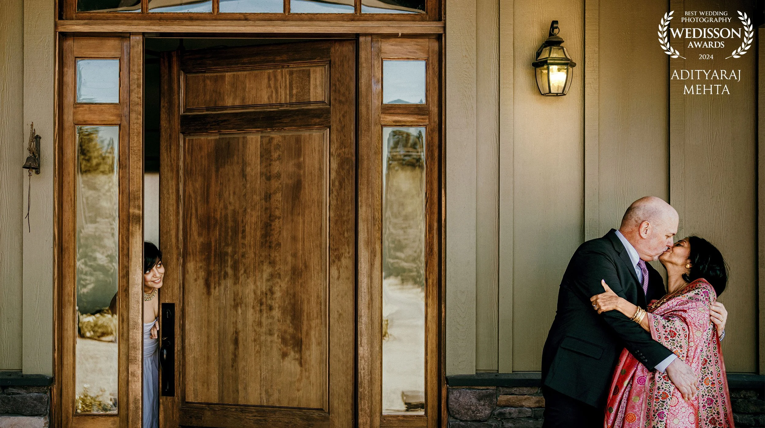 A woman in traditional attire peeking from behind a wooden door frame on the left, watching a couple sharing a kiss on the right side of the image outside the house, with a wall-mounted lantern above them.