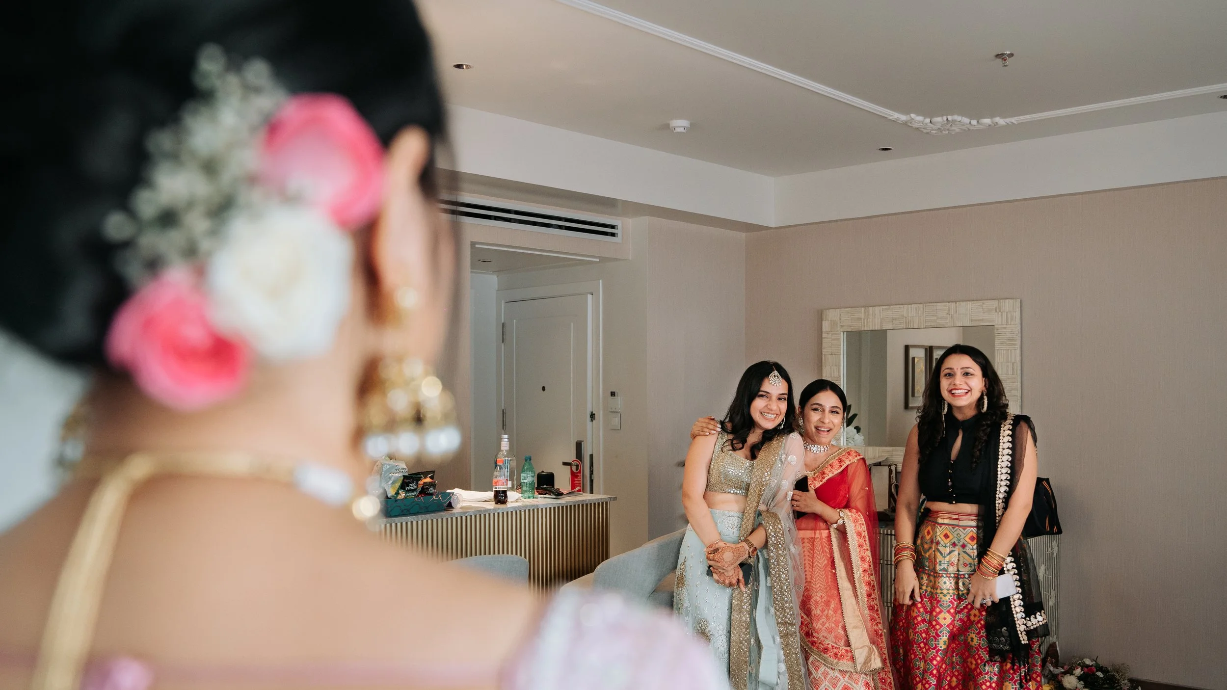 Three women in traditional Indian attire smile for a photo, while a woman with flowers in her hair is blurred in the foreground.