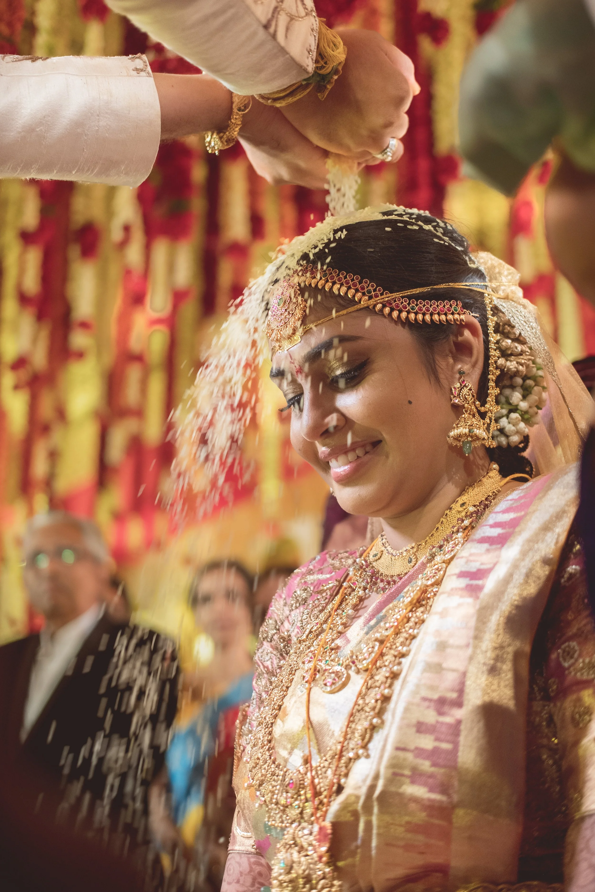 A bride dressed in traditional Indian wedding attire receives a ritual blessing with rice poured on her head during her wedding ceremony.