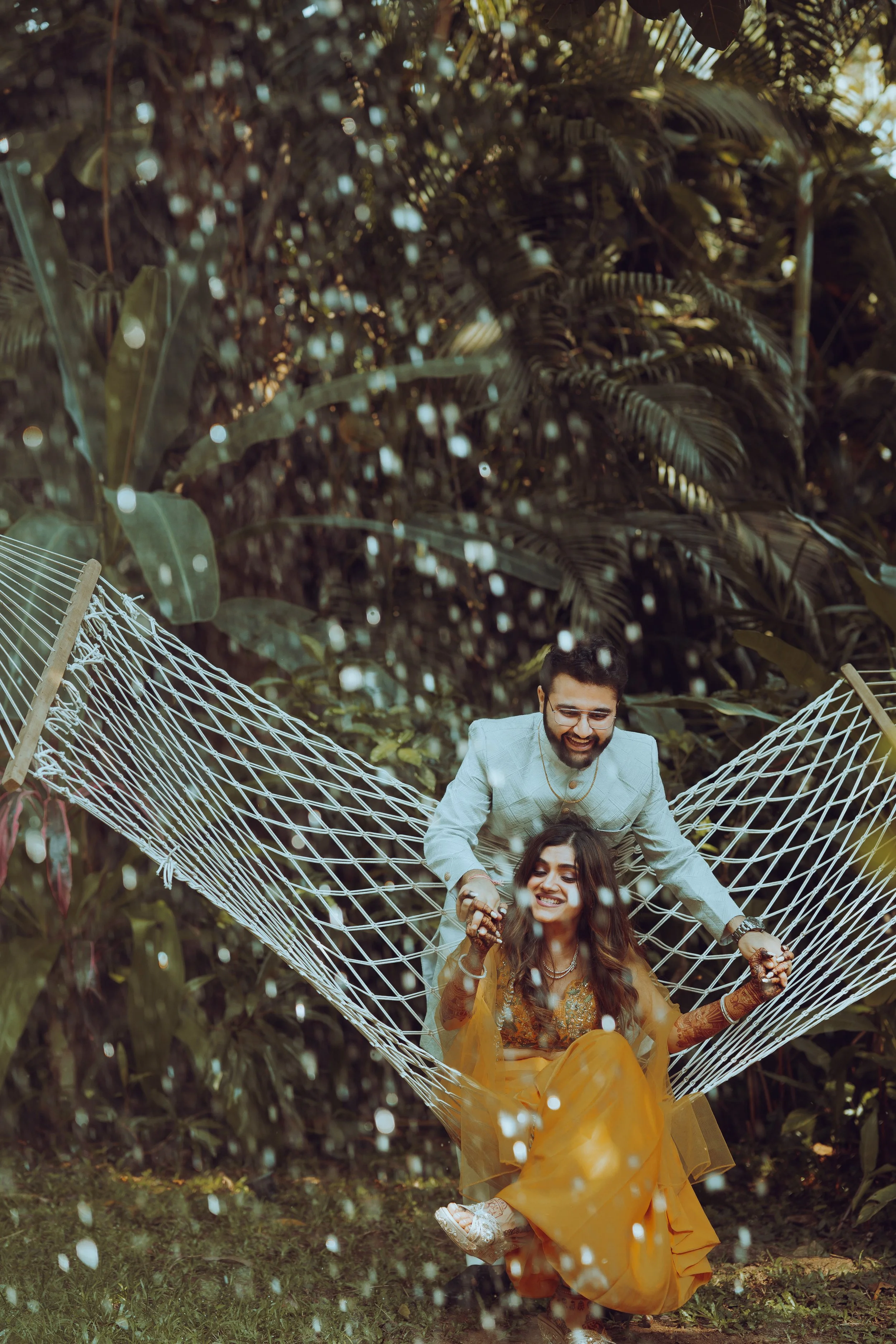 A joyful man and woman are enjoying time together in a lush garden. The woman, dressed in a vibrant yellow traditional outfit, is sitting on a hammock while the man, wearing a light-colored suit, stands behind her. They are holding hands and smiling 