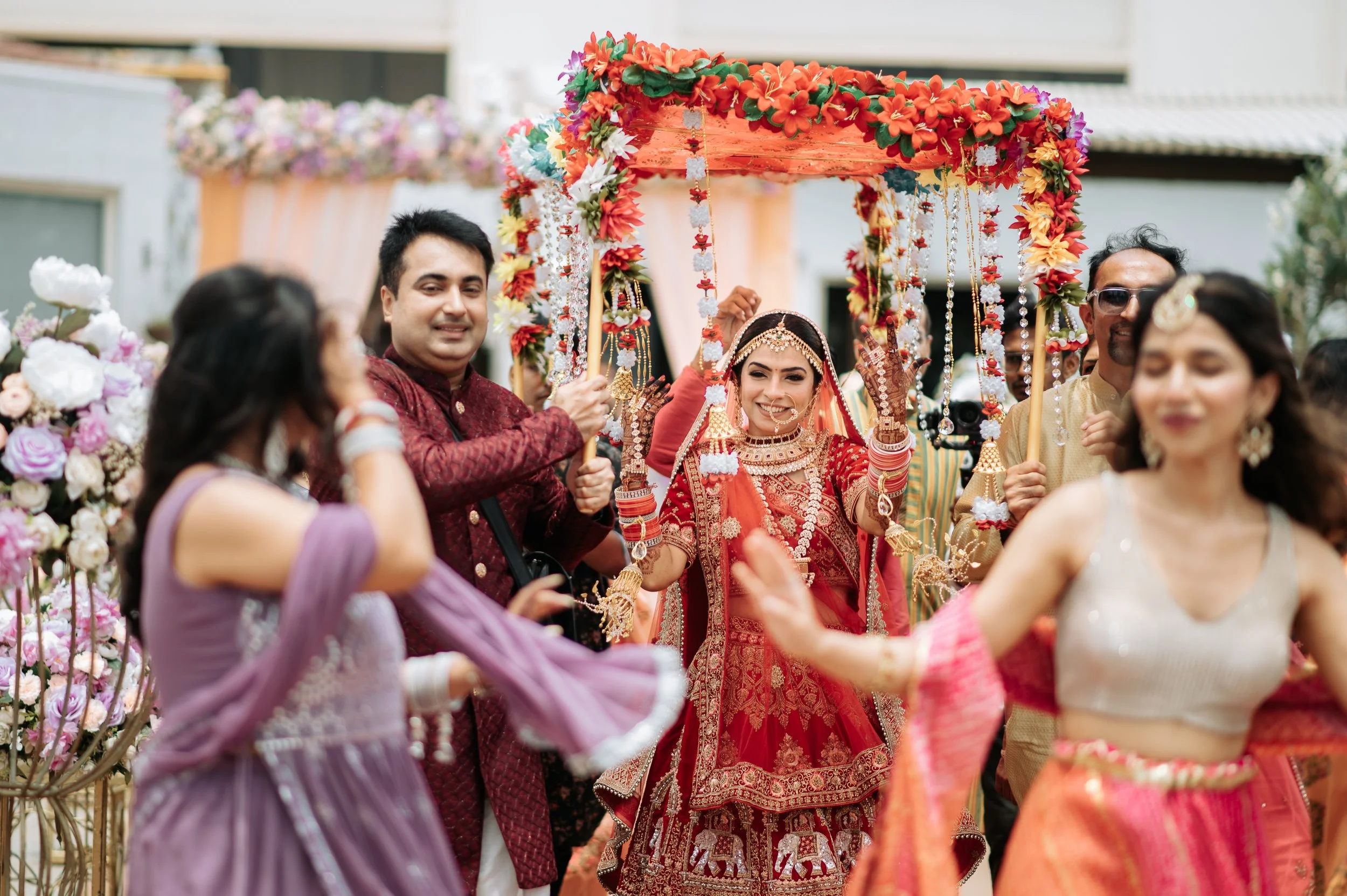 A bride in red traditional Indian wedding attire riding a decorated swing, surrounded by joyful guests at an outdoor wedding celebration.