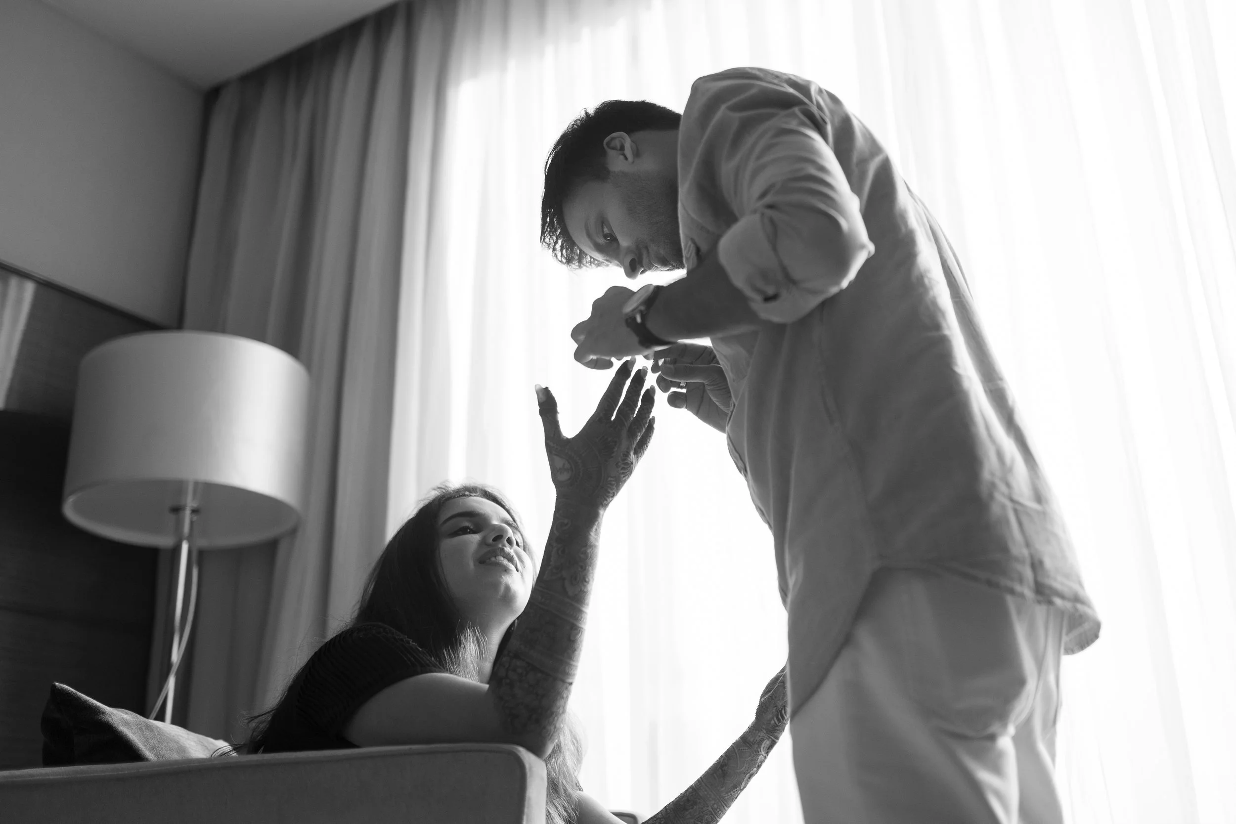 A woman with tattoos sitting on a bed, smiling, reaching up with her hand towards a man standing beside her. The man is applying a ring onto her finger. They are in a room with a curtain and a lamp in the background.