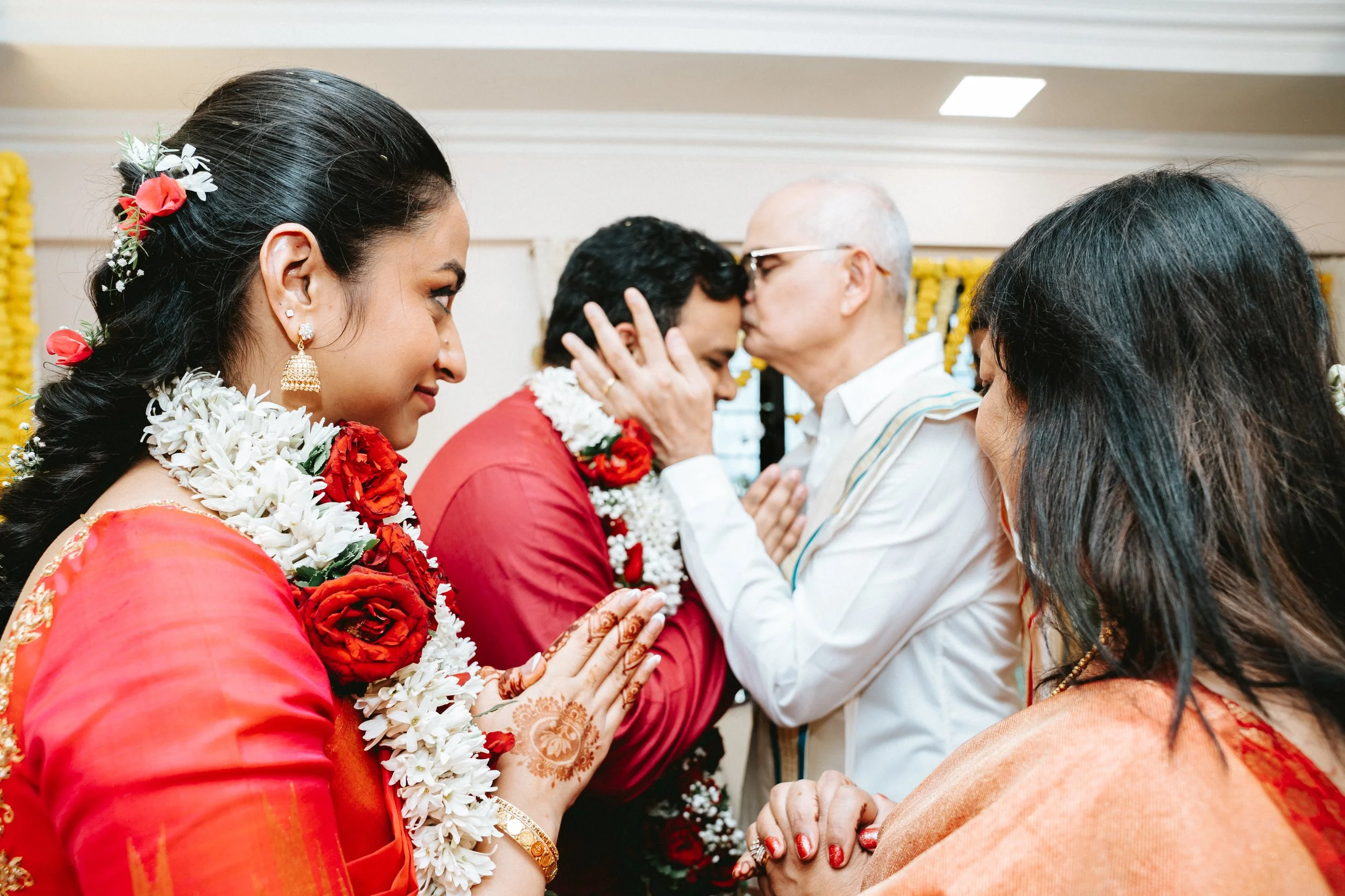 People participating in a traditional Indian wedding ceremony, with two individuals exchanging kisses and a woman with henna on her hand in the foreground.