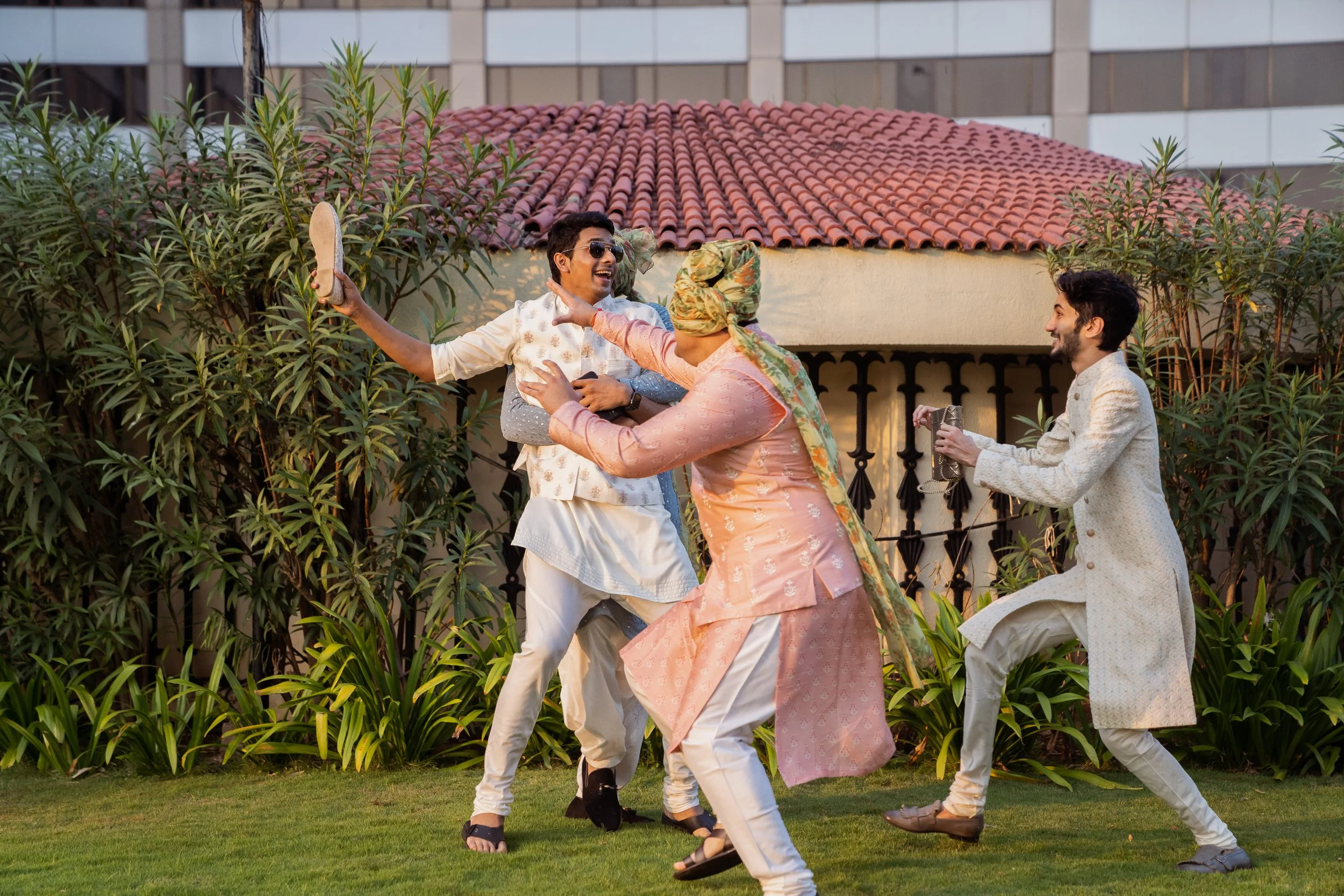Four men engaging in a joyful fight with pillows outdoors, with a small building and greenery in the background.