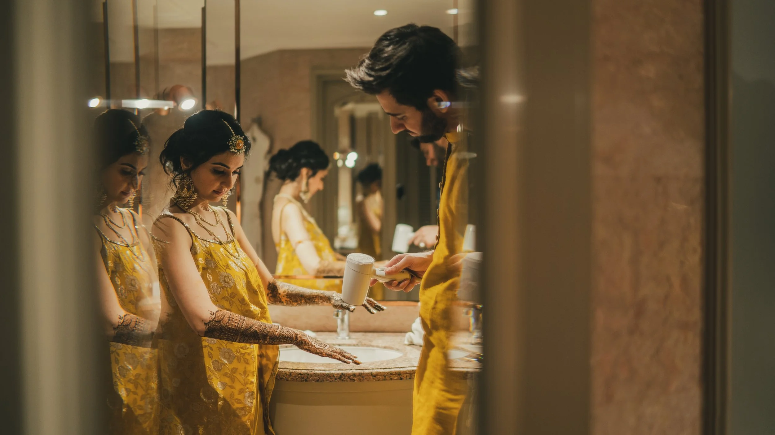 A woman in traditional Indian attire with jewelry and henna on her hands is standing at a bathroom sink, applying lotion or soap, as a man in a yellow outfit looks on and a mirror reflects her image.