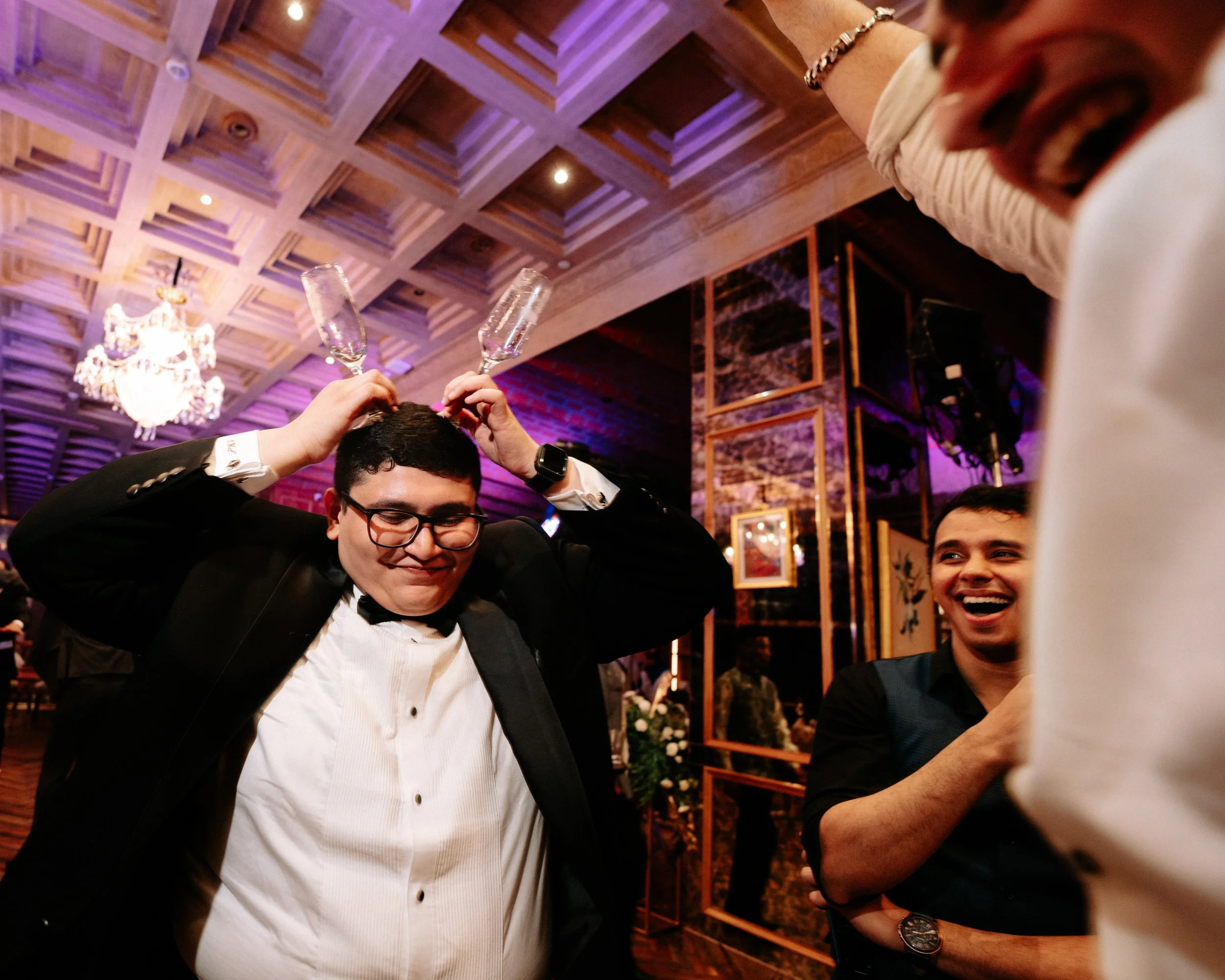 Person in formal attire balancing two empty champagne glasses on their head, surrounded by smiling friends at a lively celebration or party in a decorated venue with chandeliers and warm lighting.