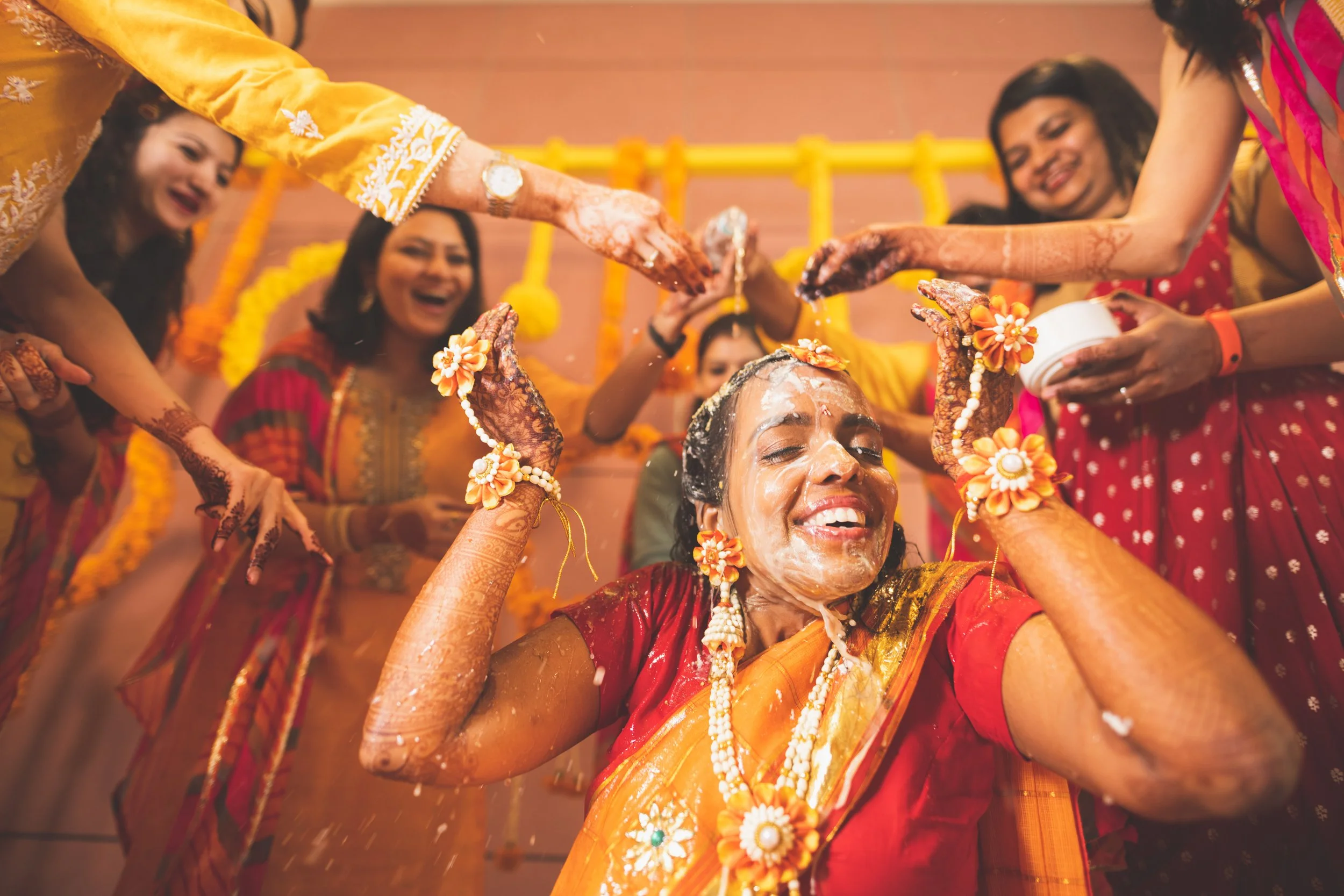 Women celebrating a traditional Indian wedding with a henna ceremony, applying turmeric on bride's face, wearing colorful sarees, and decorated with flowers.