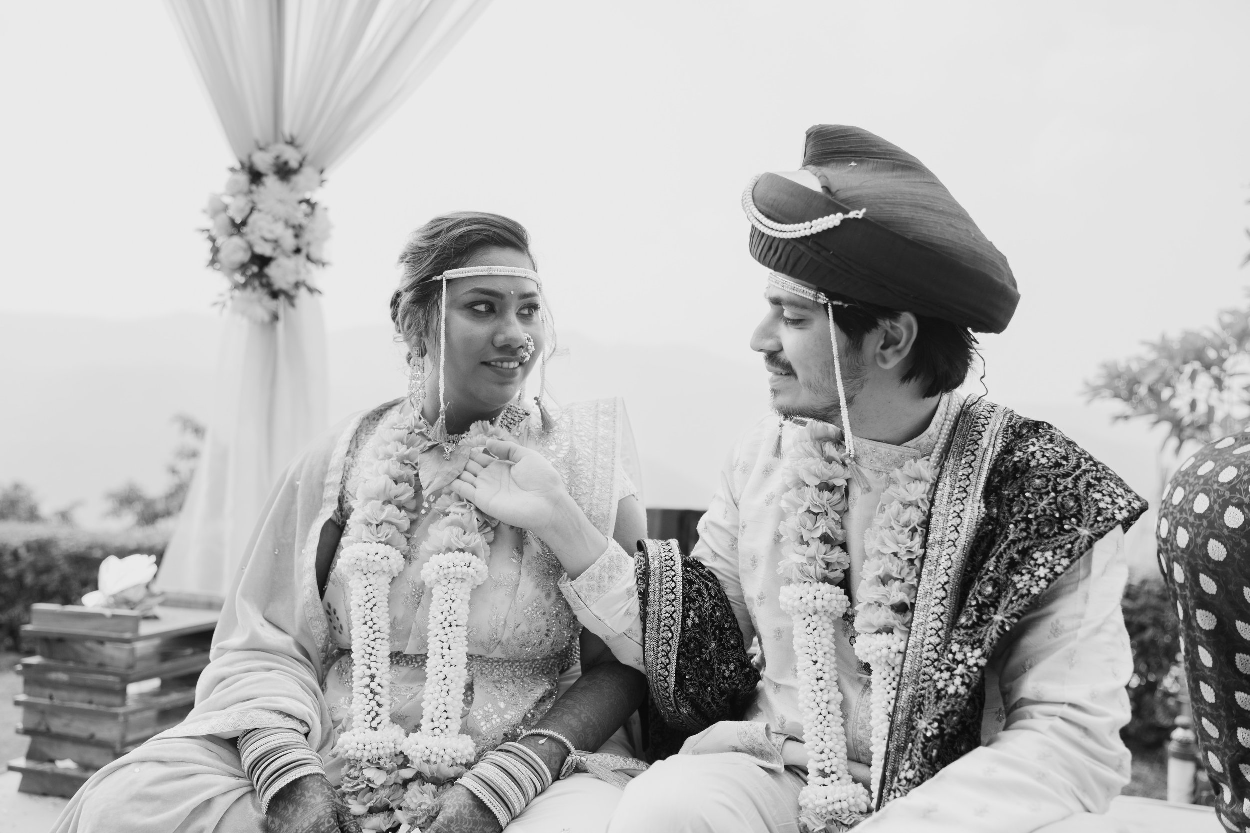 Black and white photo of a bride and groom during a traditional Indian wedding ceremony. The bride is wearing traditional attire and jewelry, with floral garlands, while the groom is wearing a turban and a decorated shawl. They are sitting outdoors u