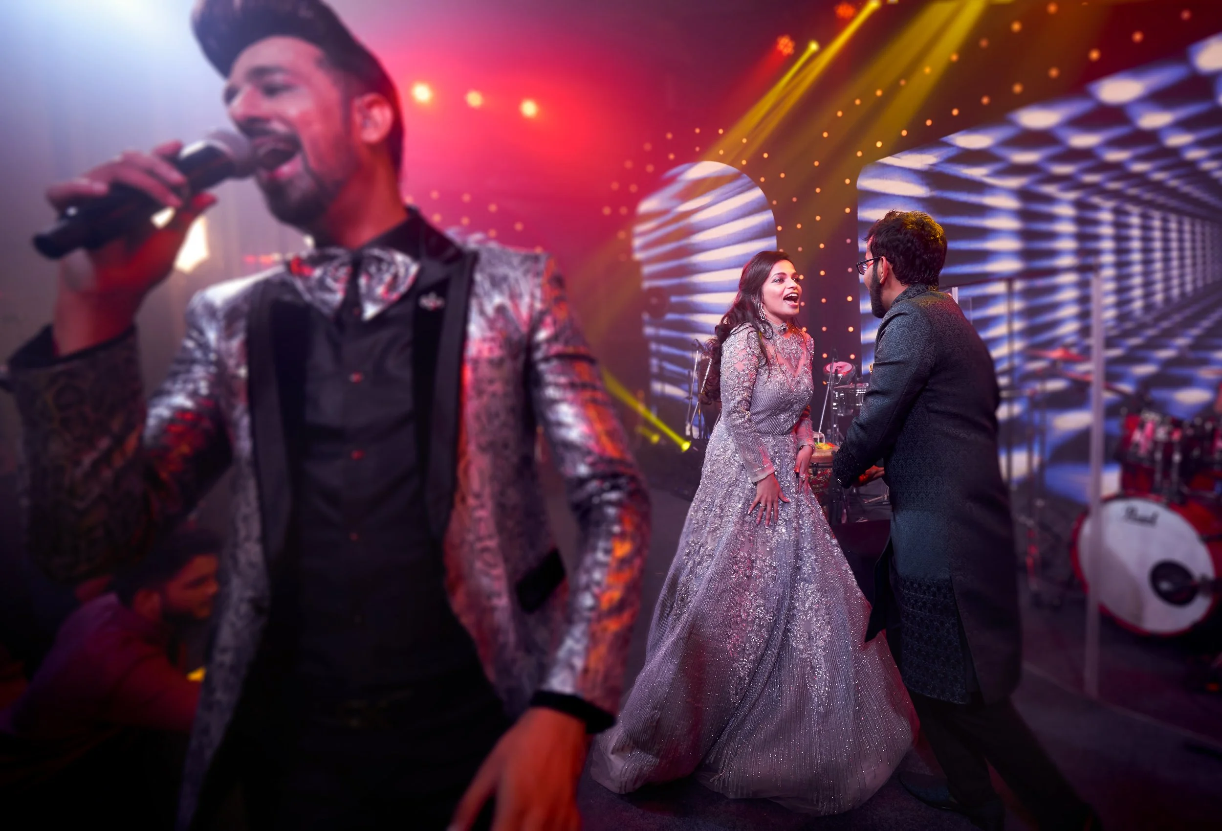 People singing and dancing at a party with colorful lights and a stage backdrop.