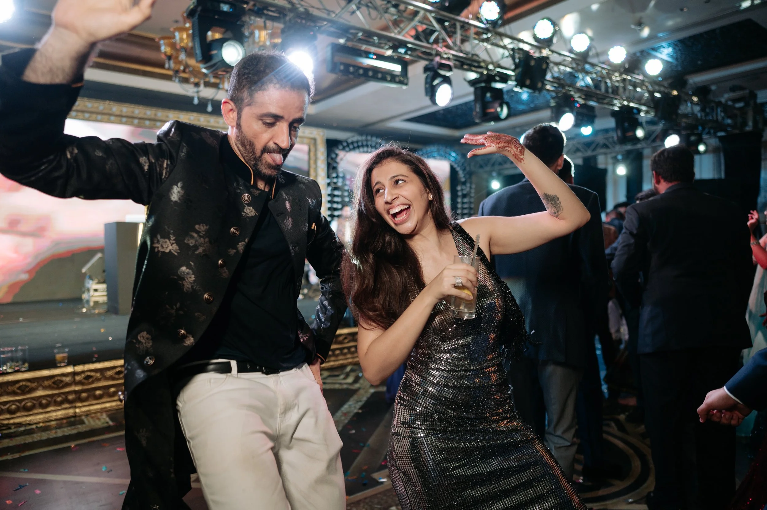 Two people dancing and enjoying a celebration at a party. The man is wearing a black jacket with floral patterns and white pants, and the woman is wearing a shiny dress and holding a drink. The background shows a decorated event space with other gues