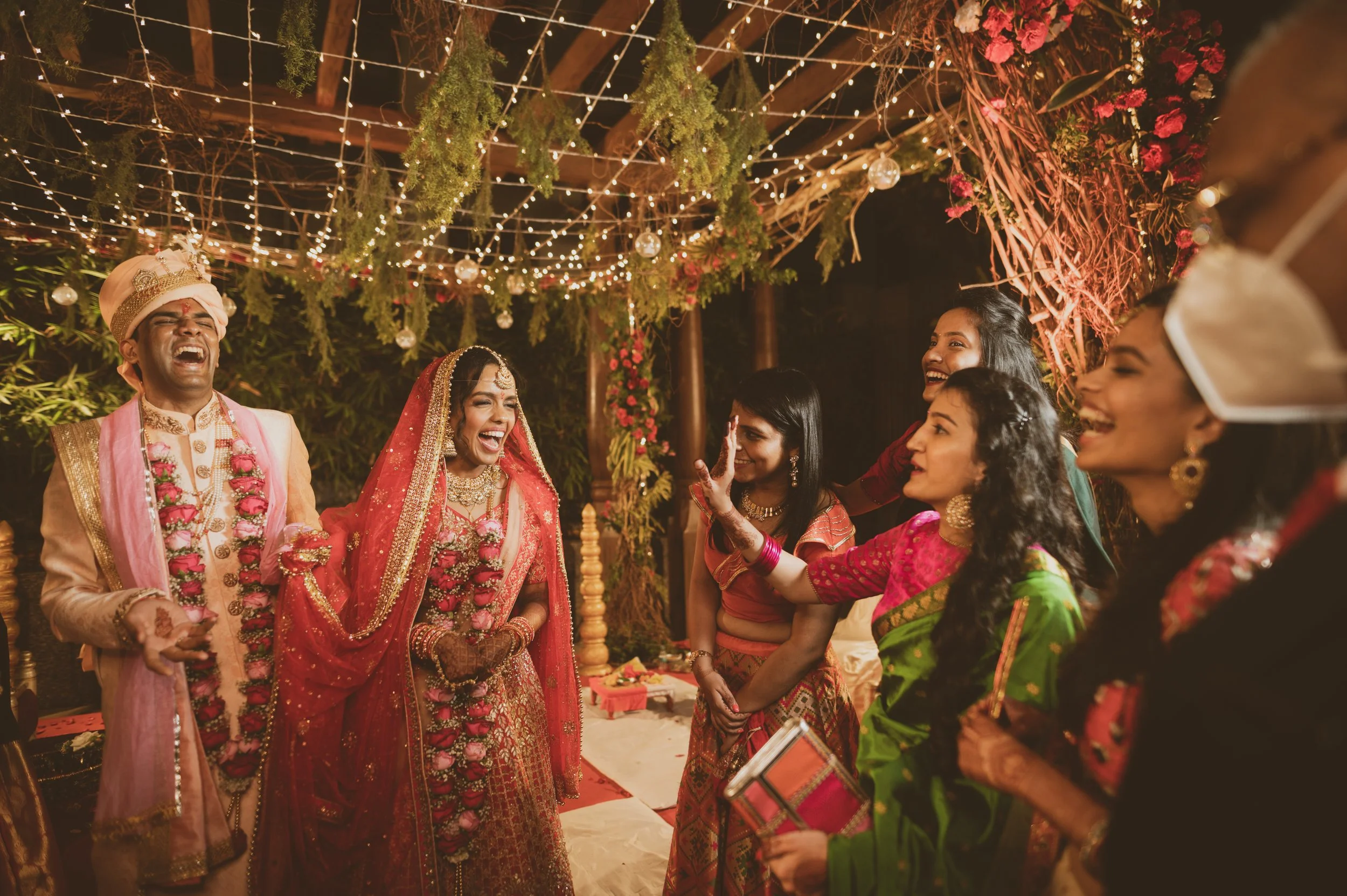 A joyful Indian bride and groom in traditional wedding attire laughing with friends and family under string lights and floral decorations at night.