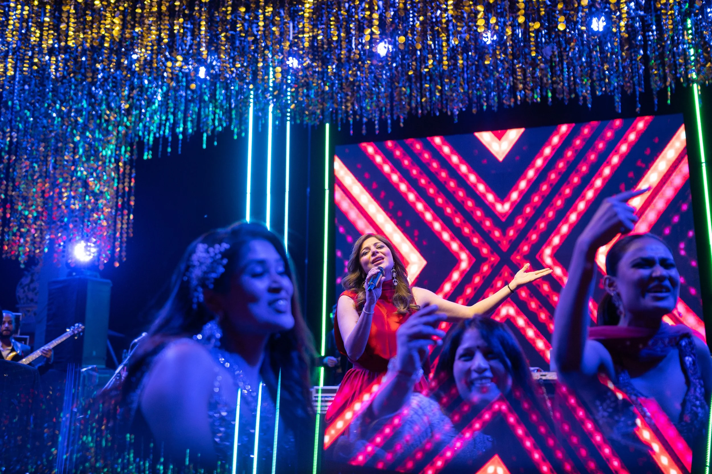 A group of women singing on stage with colorful, bright lights and a digital backdrop, with a musician visible playing guitar in the background.