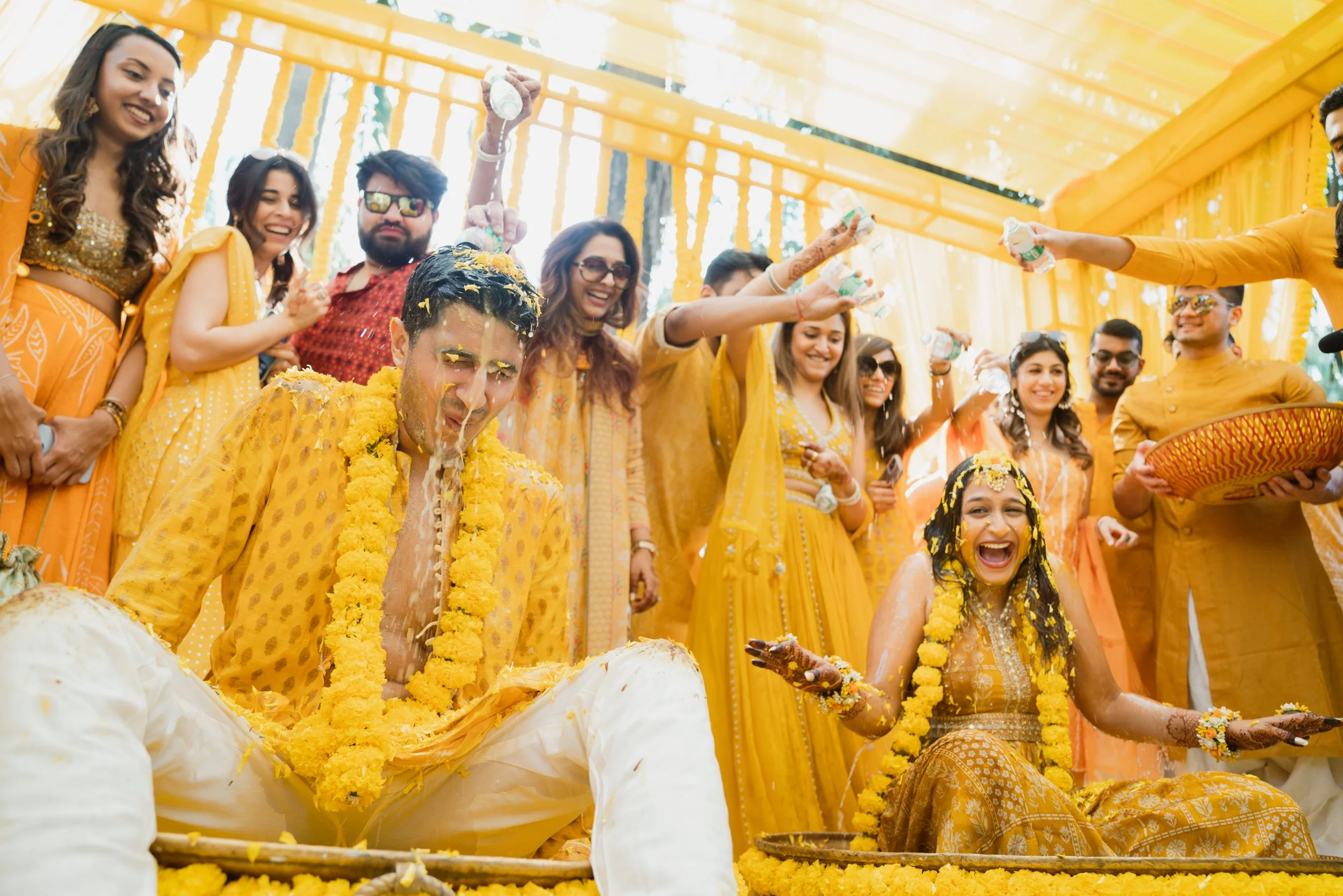 Indian wedding celebration with the bride and groom covered in yellow flower garlands, surrounded by family and friends in yellow attire, participating in a traditional ceremony involving pouring and throwing rice or flower petals.