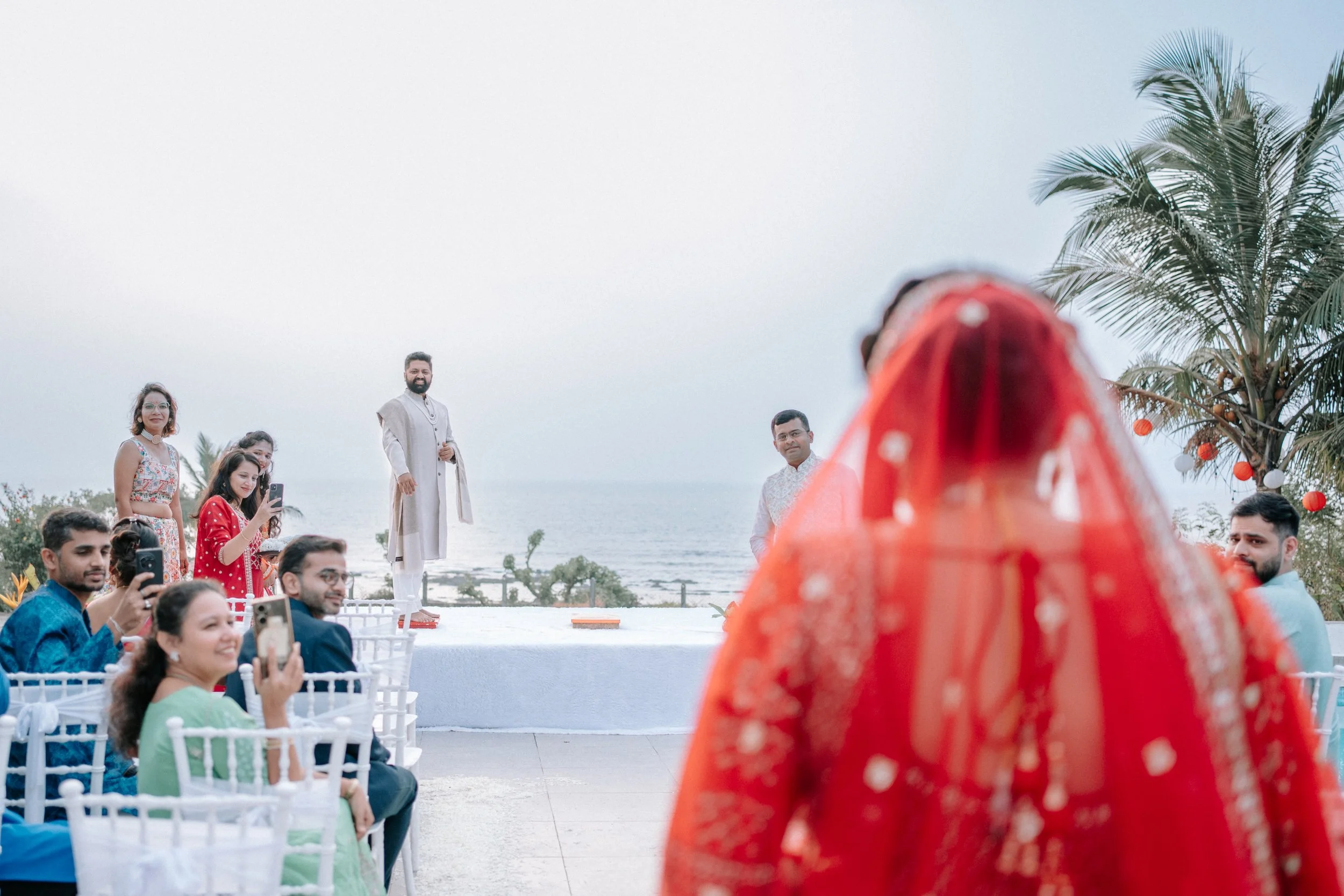A wedding ceremony taking place outdoors near the ocean with guests seated in white chairs. In the foreground, a bride in a red dress with gold embellishments is facing the guests. In the background, a groom and officiant are standing on a white plat