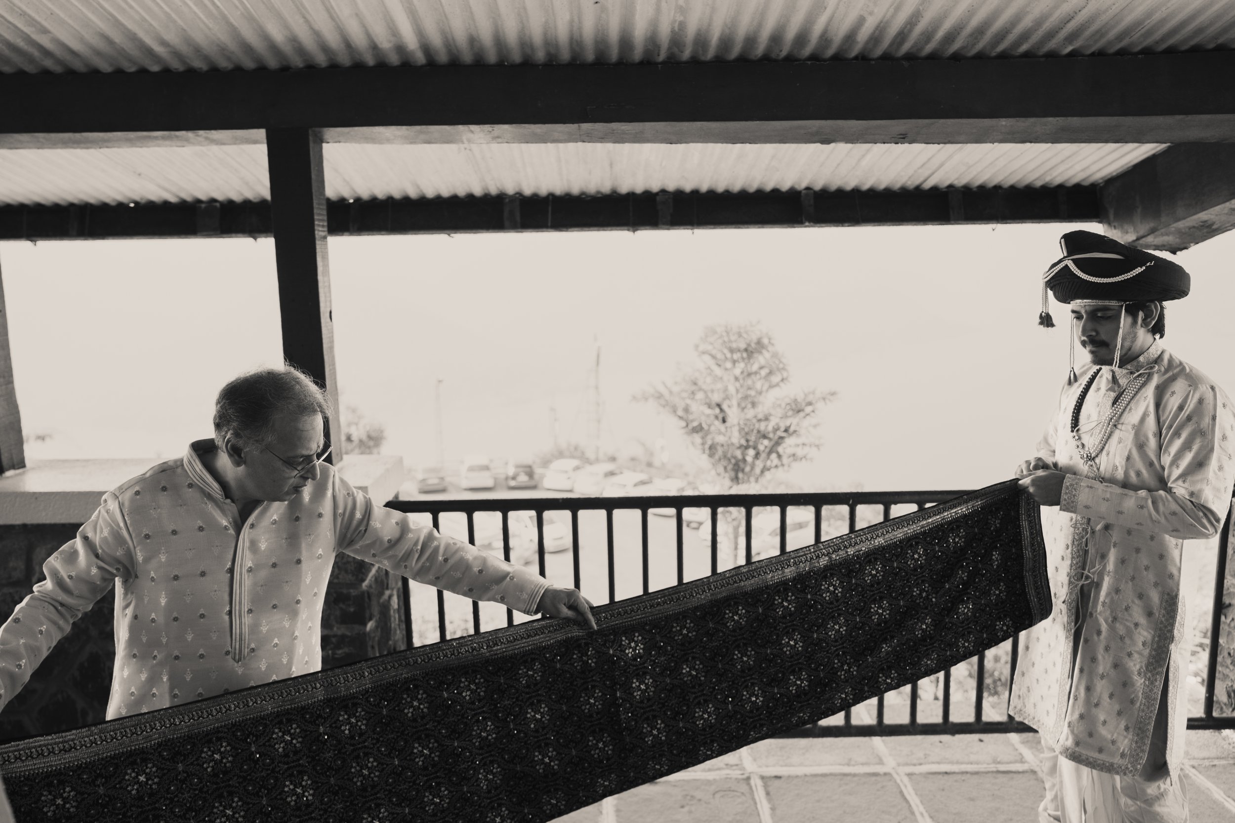 Two men wearing traditional Indian attire holding a decorative rug on a balcony with a parking lot and trees in the background, in black and white.
