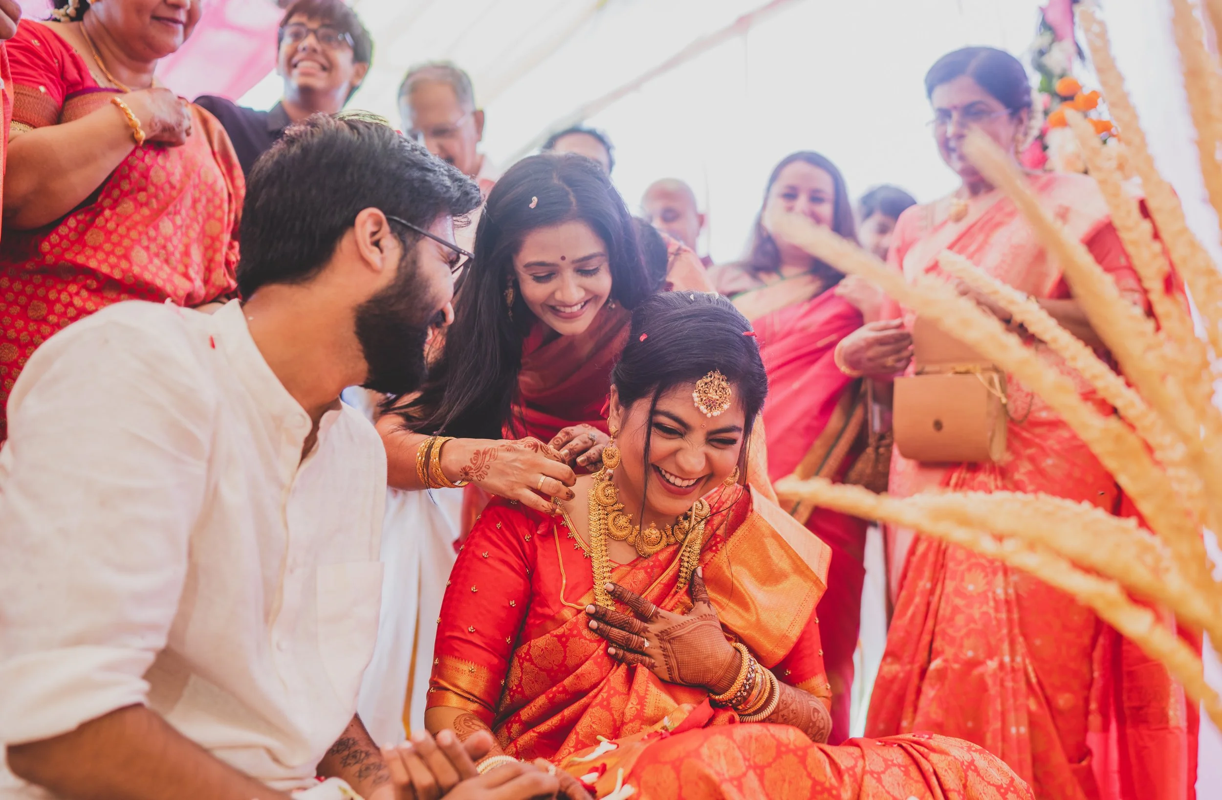 Indian wedding ceremony with a bride in a red and gold sari, surrounded by guests, some dressed in traditional sarees, participating in a ritual and smiling.