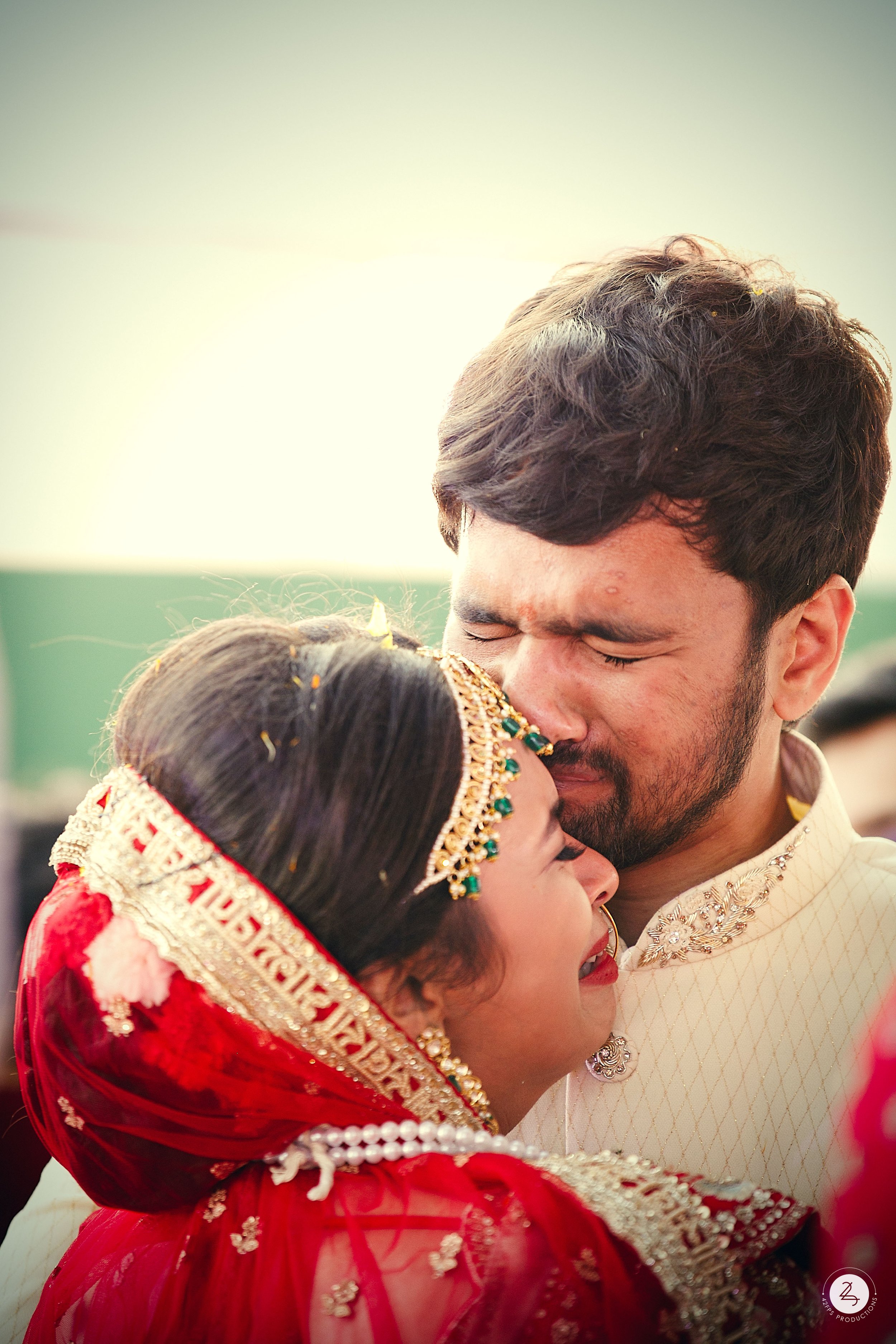 A couple in traditional Indian wedding attire sharing a close, emotional moment, with the woman in a red saree and jewelry, and the man in a cream-colored kurta.