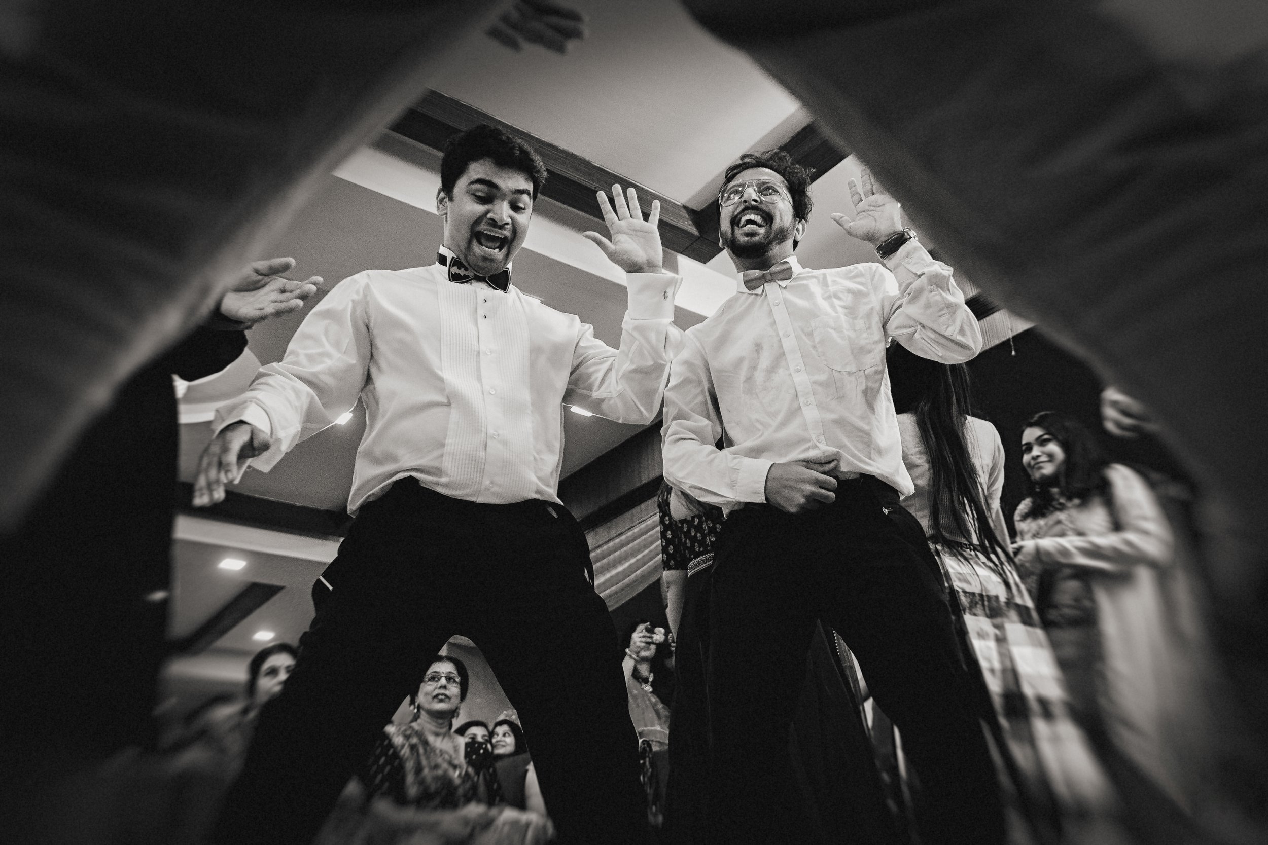 Two men in formal attire dancing and cheering at a celebration or party, viewed from beneath a table or low vantage point.