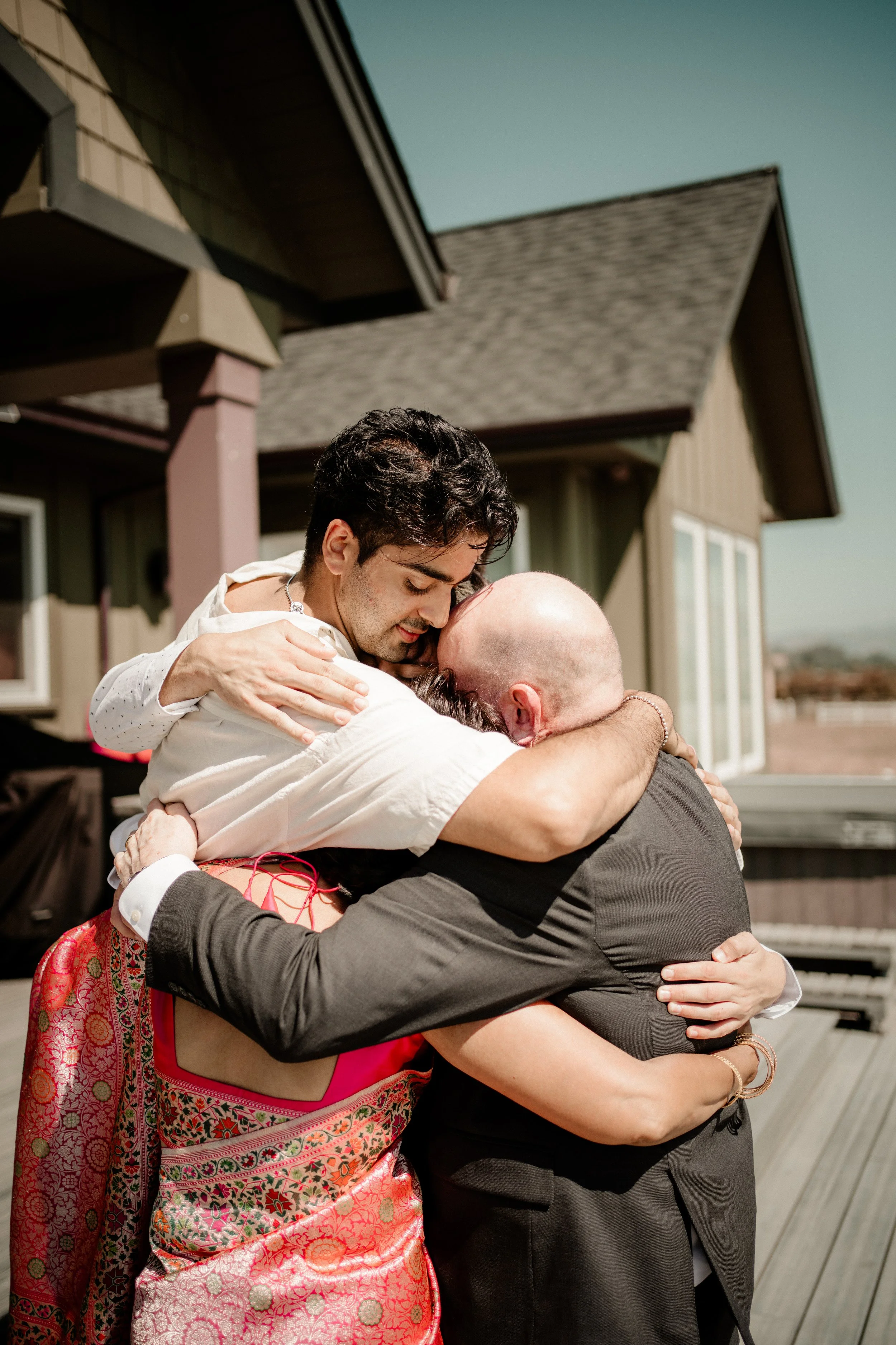 Three people embracing and hugging each other outside a house on a sunny day.