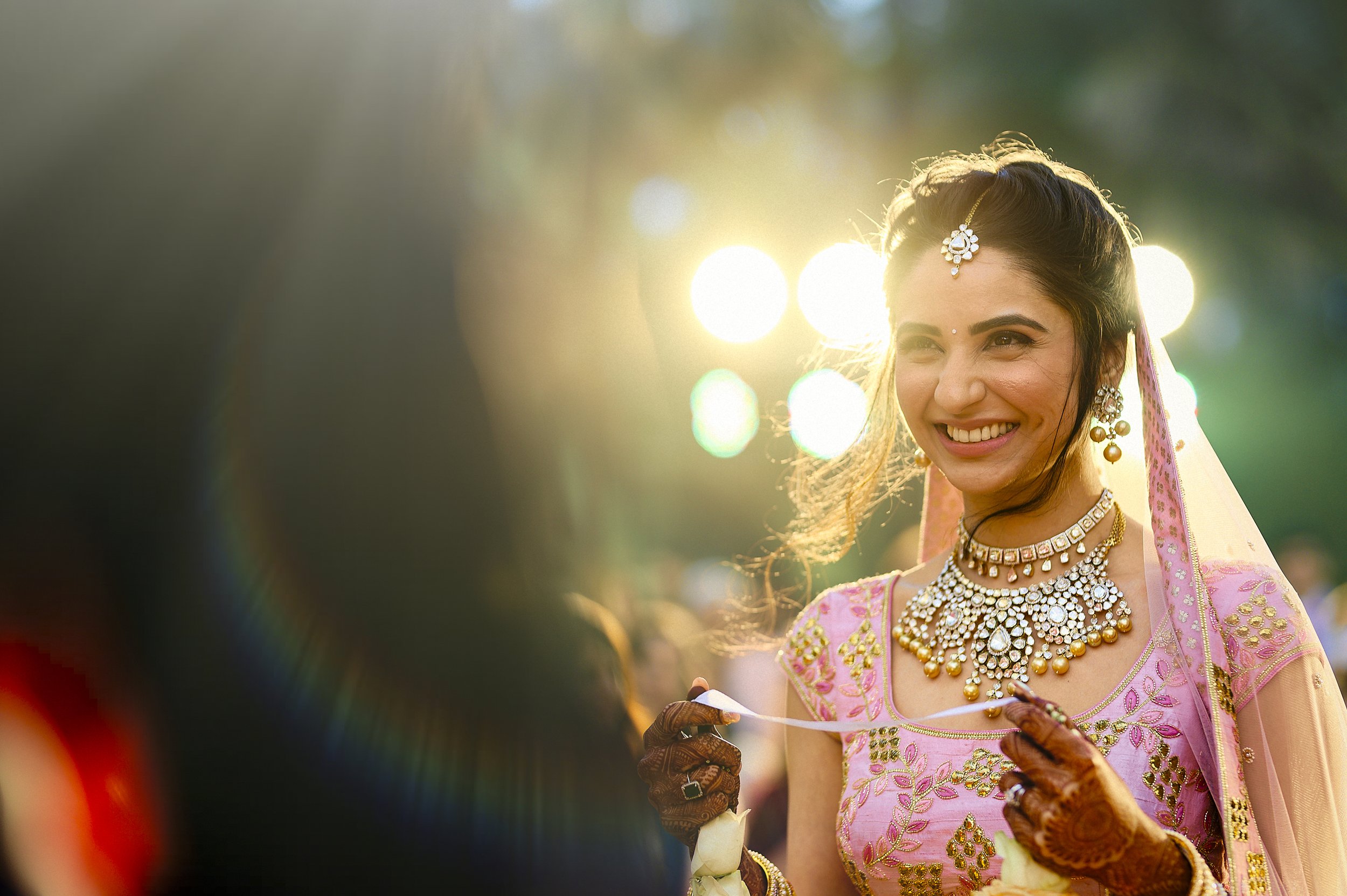 A woman dressed in traditional Indian jewelry and pink saree smiling at a wedding celebration outdoors with sunlight in the background.