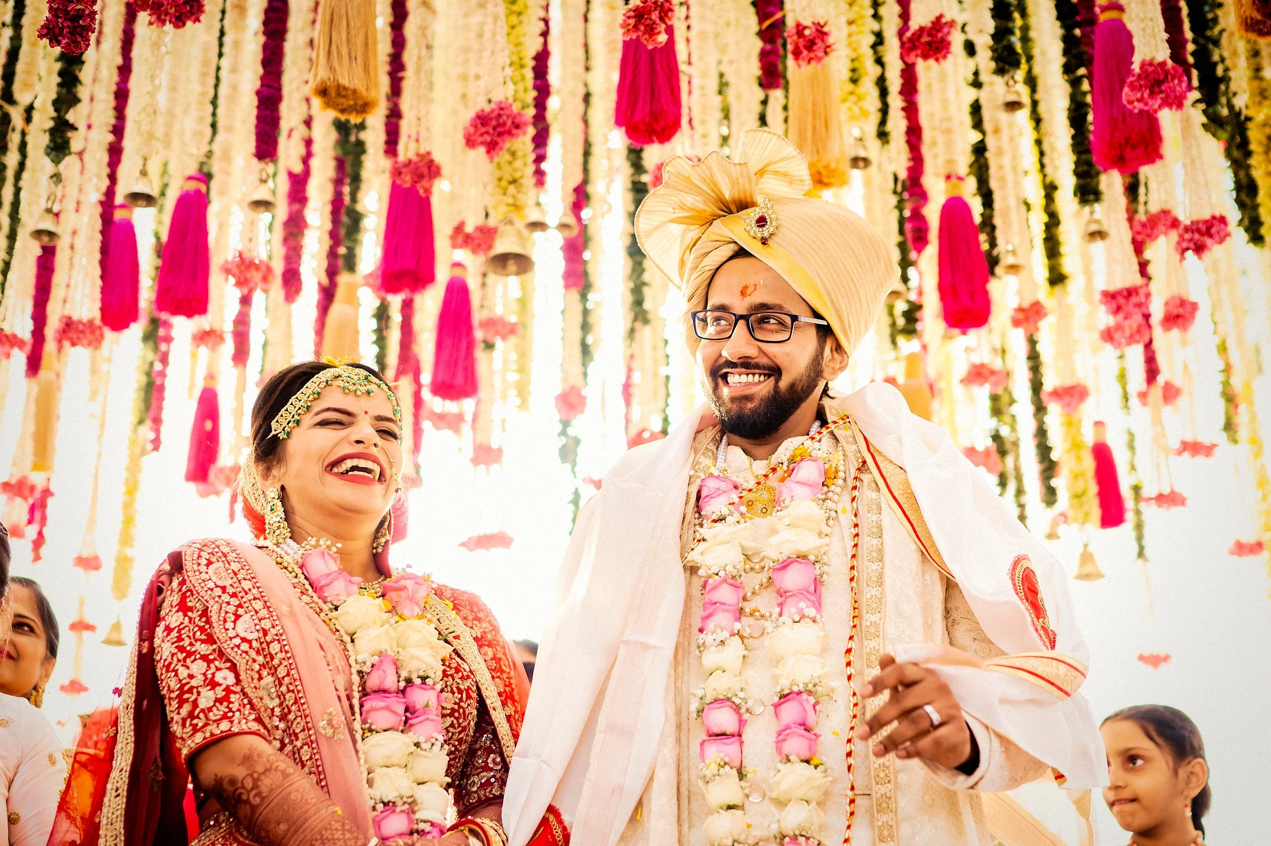A joyful Indian wedding ceremony with the bride and groom in traditional attire, wearing garlands, under colorful hanging floral decorations.
