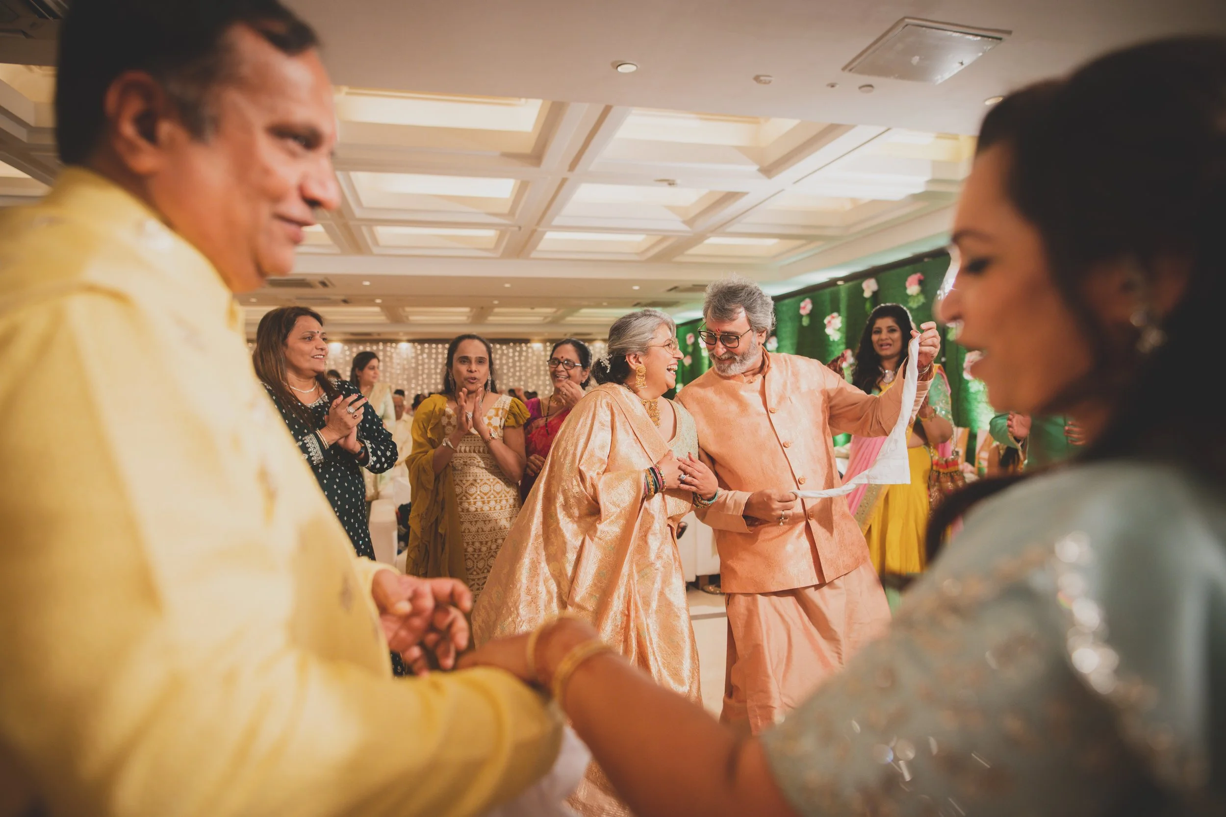 Indian wedding celebration with guests dancing and interacting, featuring a woman in a gold saree and a man in peach traditional attire sharing a joyful moment in a decorated hall.