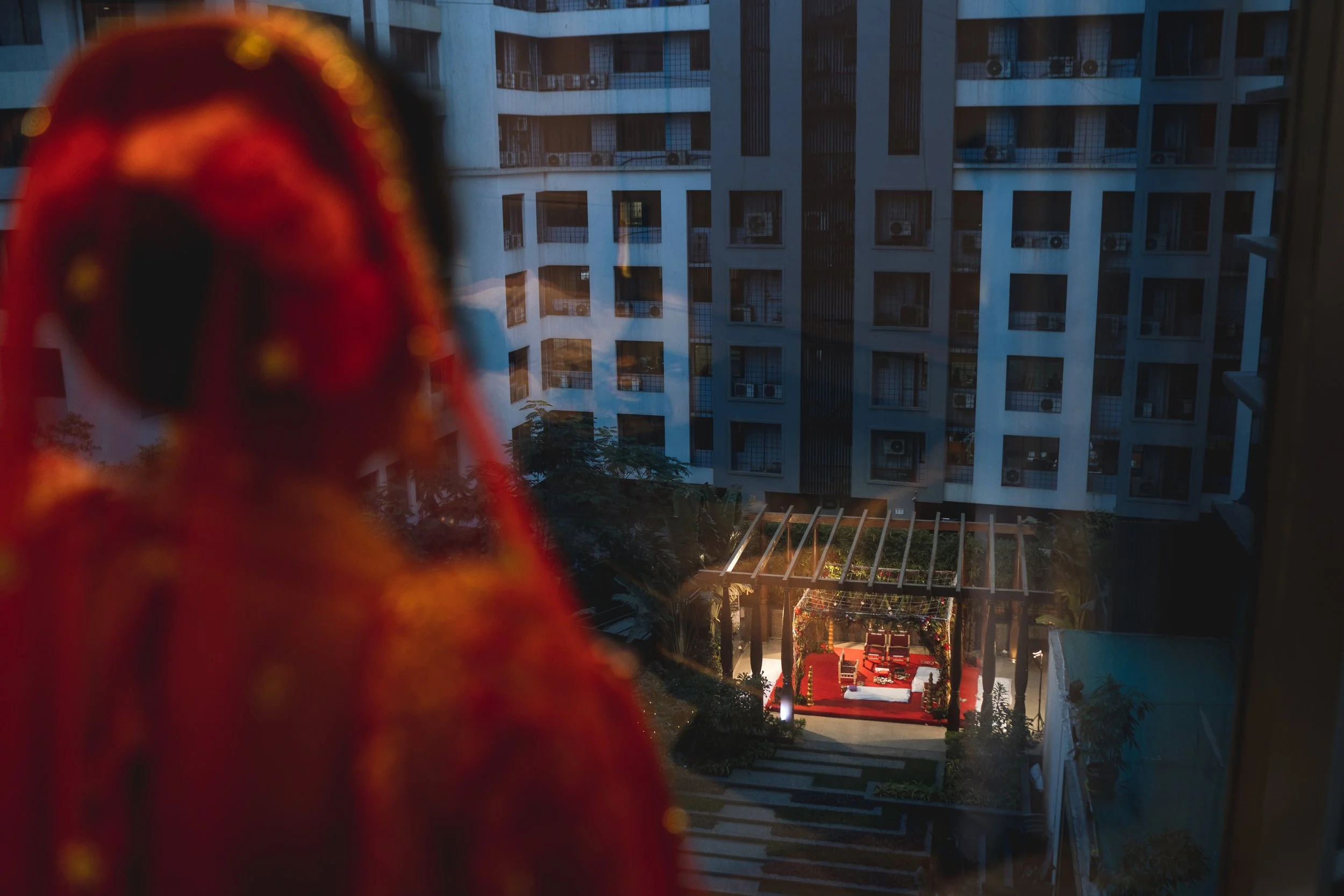 A blurry figure draped in red standing in front of a window, with a view of an outdoor courtyard and modern apartment buildings at dusk.